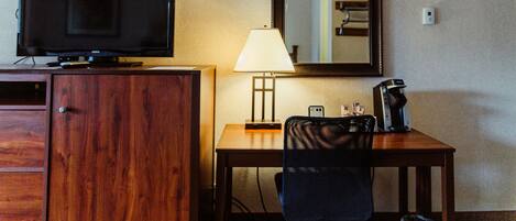 A hotel room with a wooden desk, a television, a lamp, a coffee maker, and a mirror on the wall.