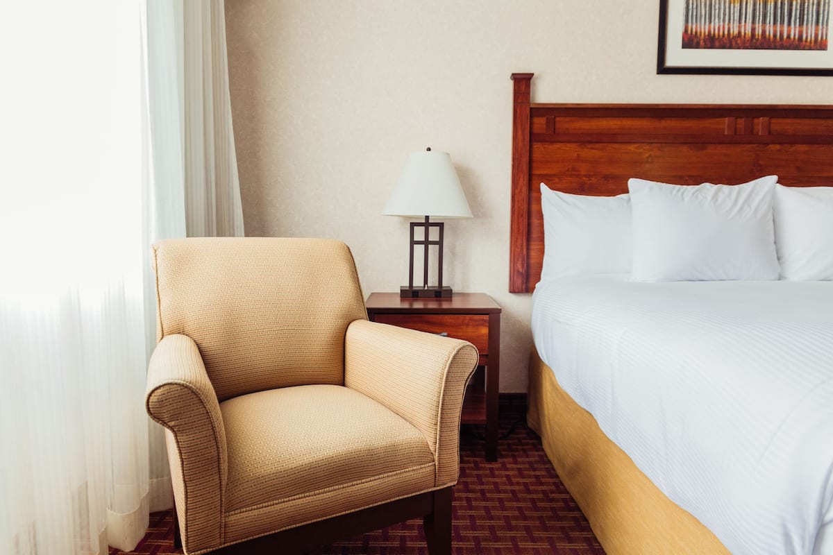 A hotel room with a bed, a chair, a nightstand with a lamp, and a window with curtains.