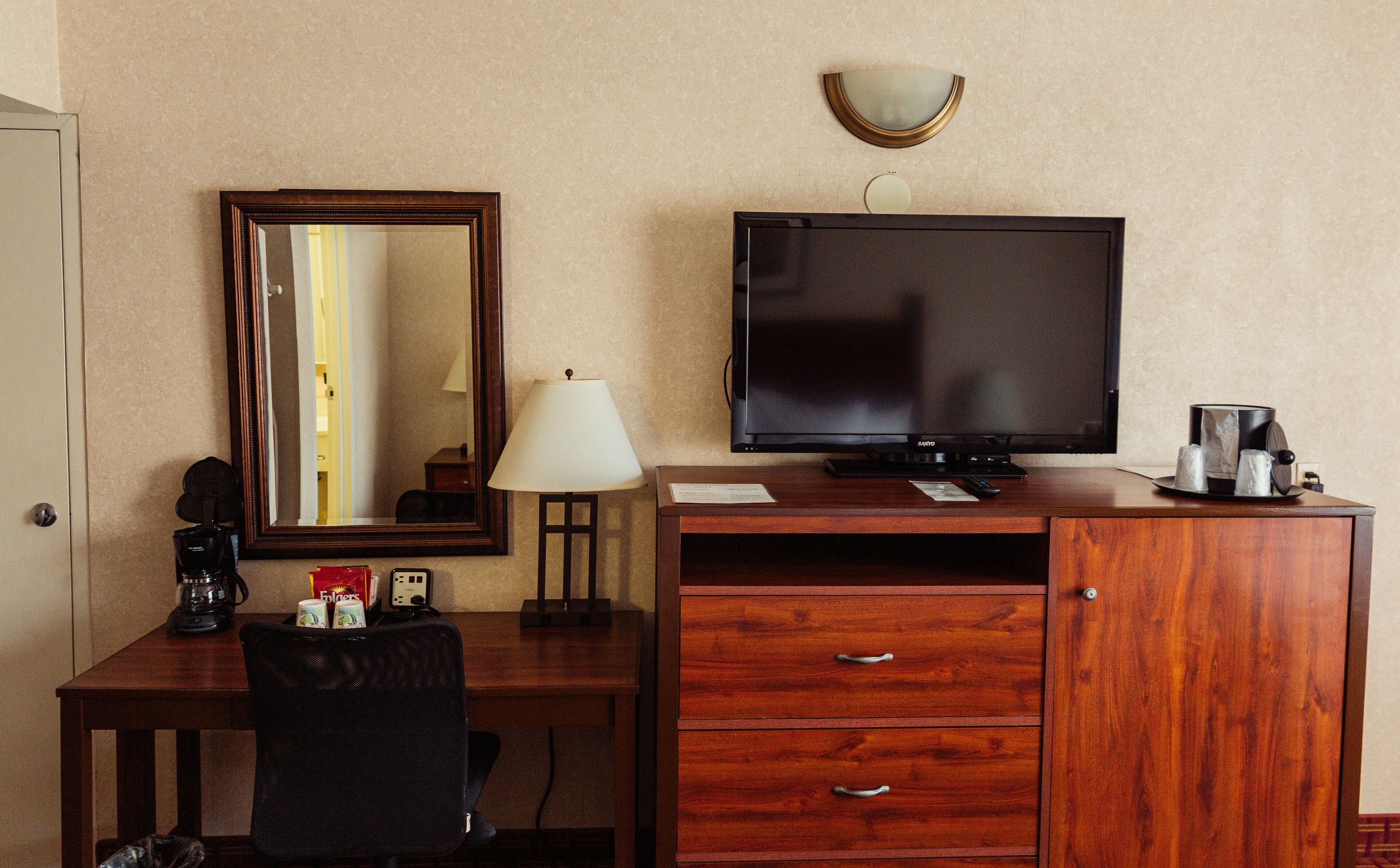 A hotel room with a wooden desk, a television, a mirror, a lamp, and a minibar.