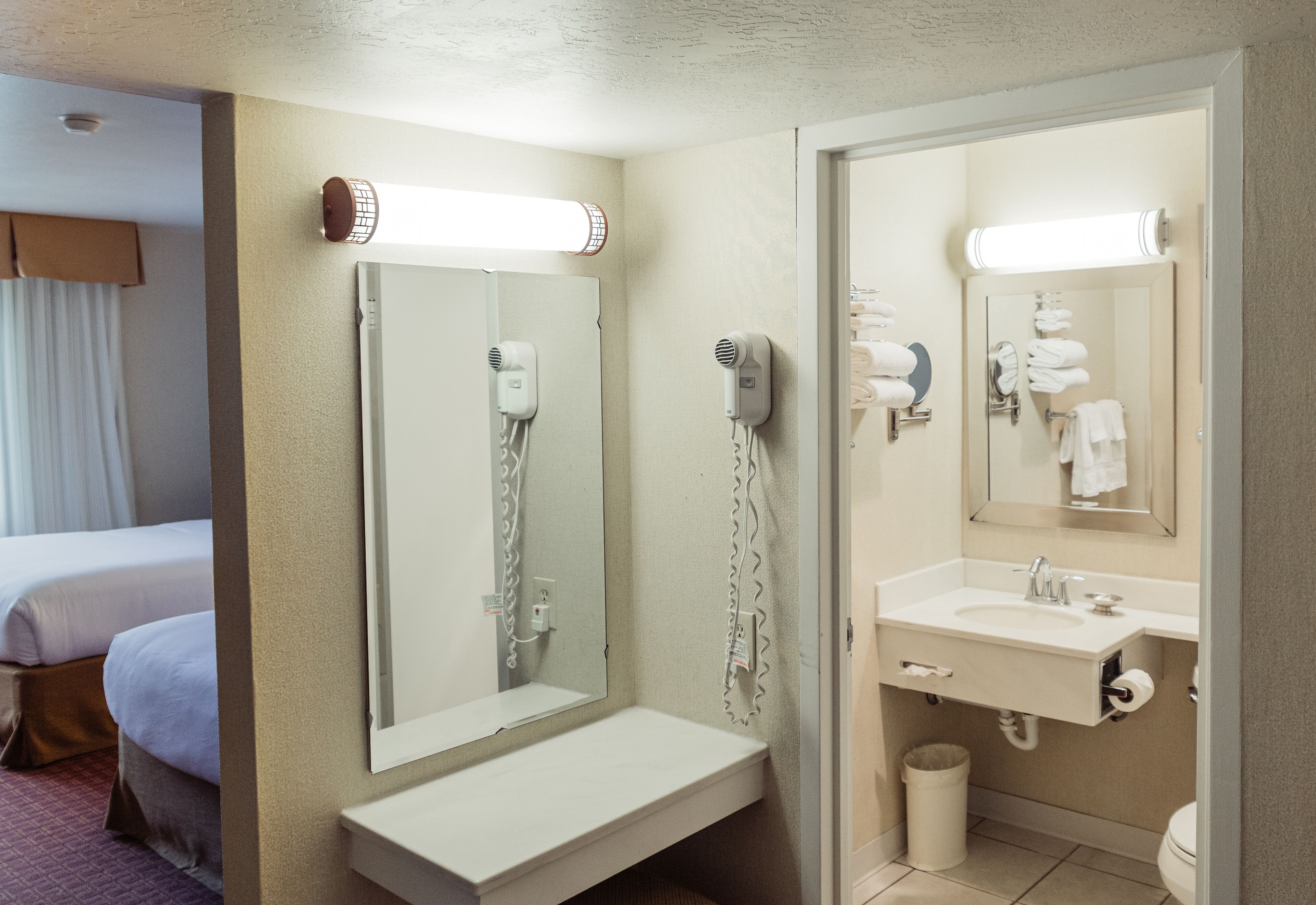 A hotel bathroom with a large mirror, a hairdryer, a sink, and a toilet.