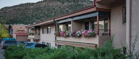 A building with a balcony adorned with flowers, parked cars, and a mountainous backdrop.
