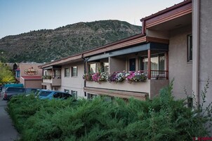 A building with a balcony adorned with flowers, parked cars, and a mountainous backdrop.