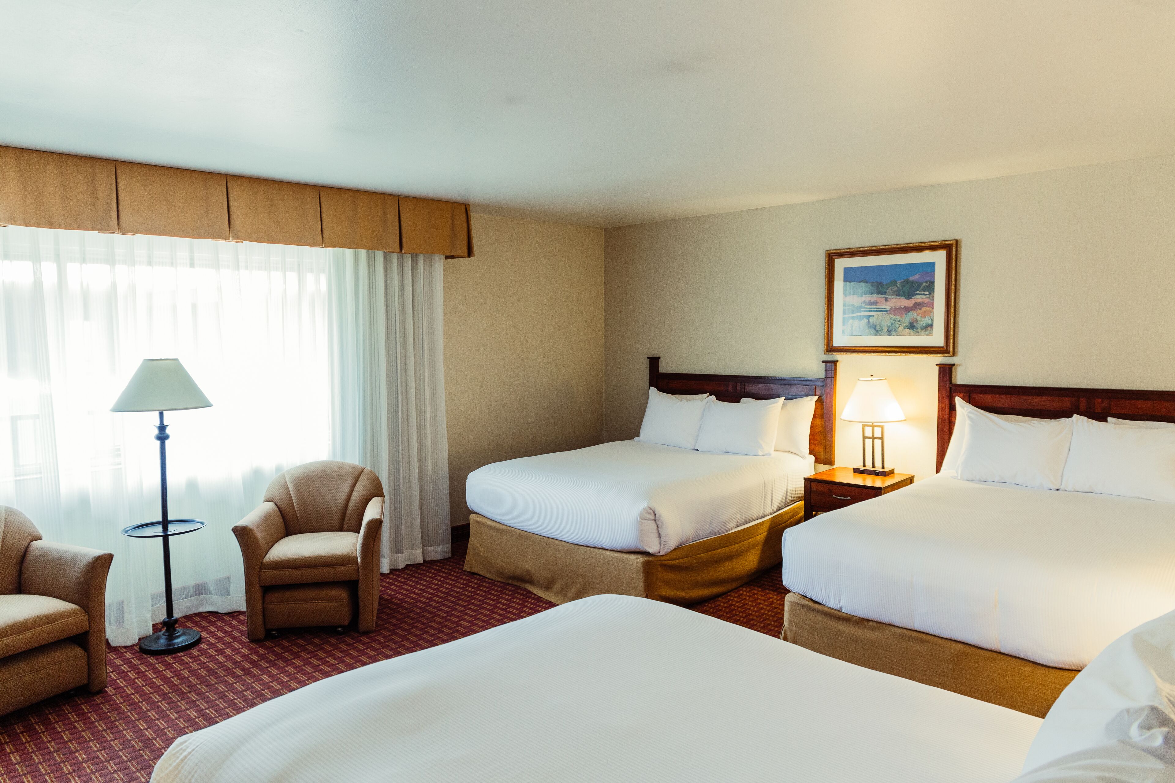 A hotel room with two beds, a chair, a lamp, and a painting on the wall.