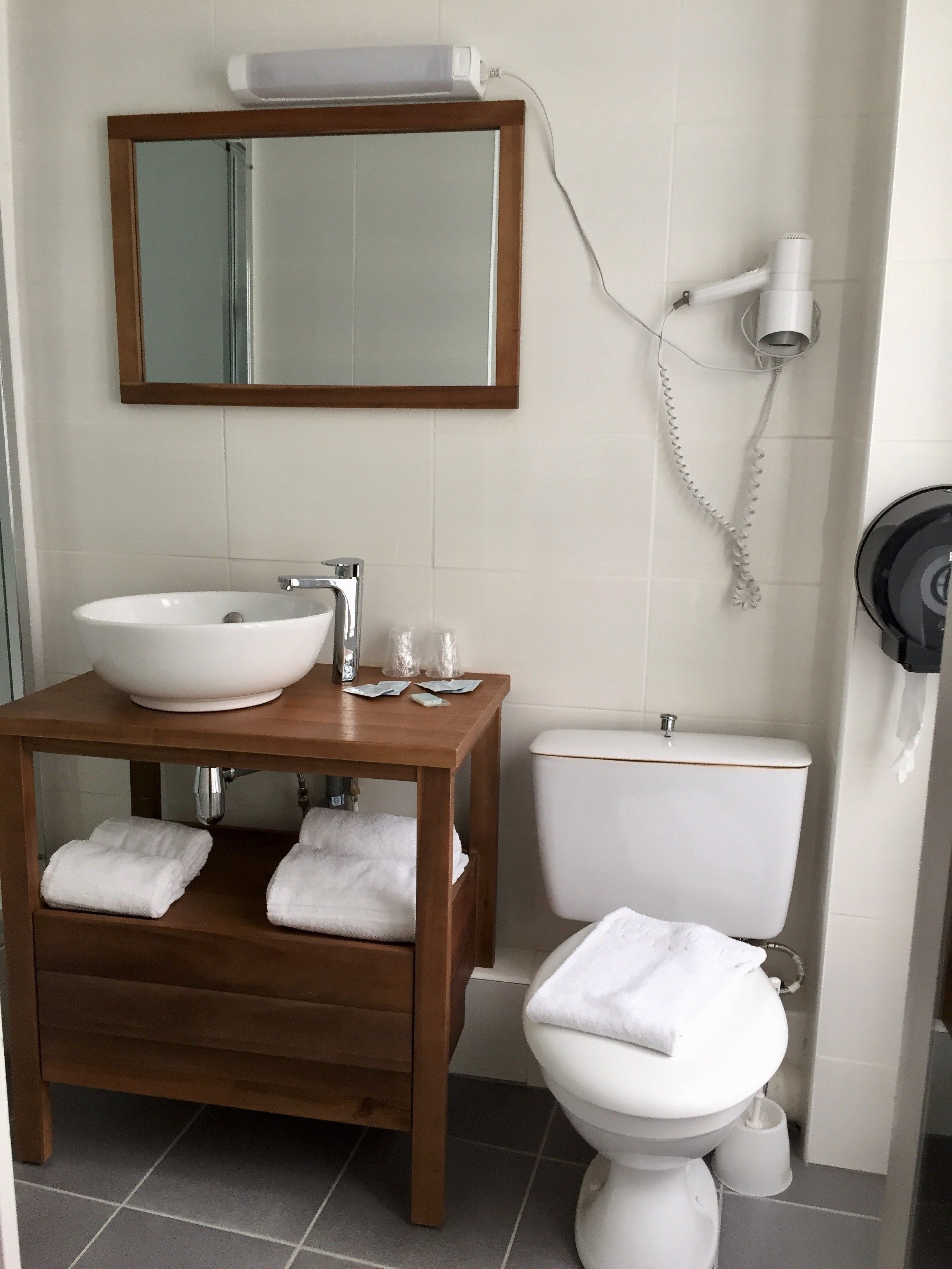 A bathroom with a wooden vanity, a white sink, a mirror, a toilet, and a hairdryer on the wall.