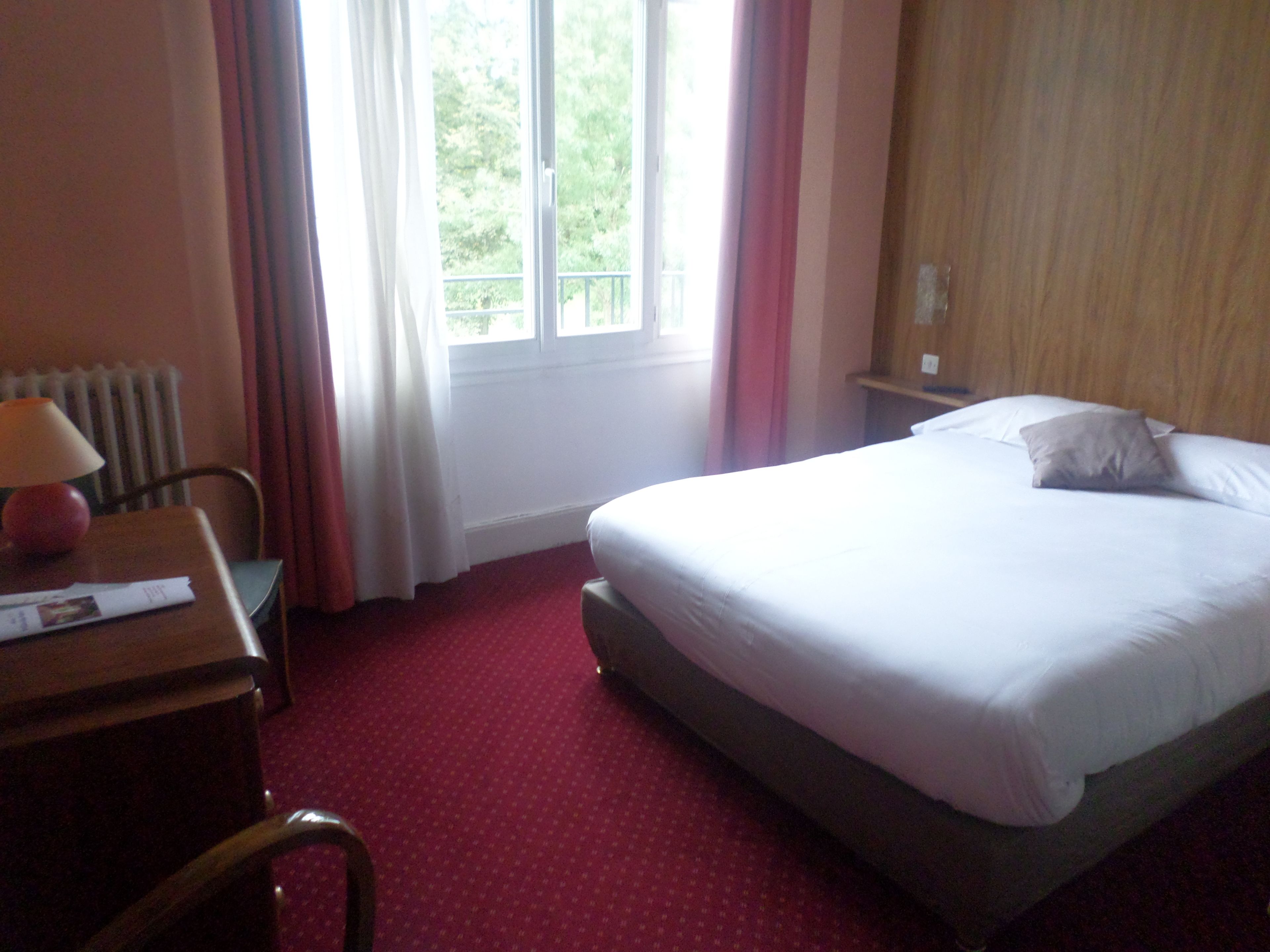 A hotel room with a bed, a desk with a lamp, a chair, and a window with curtains.