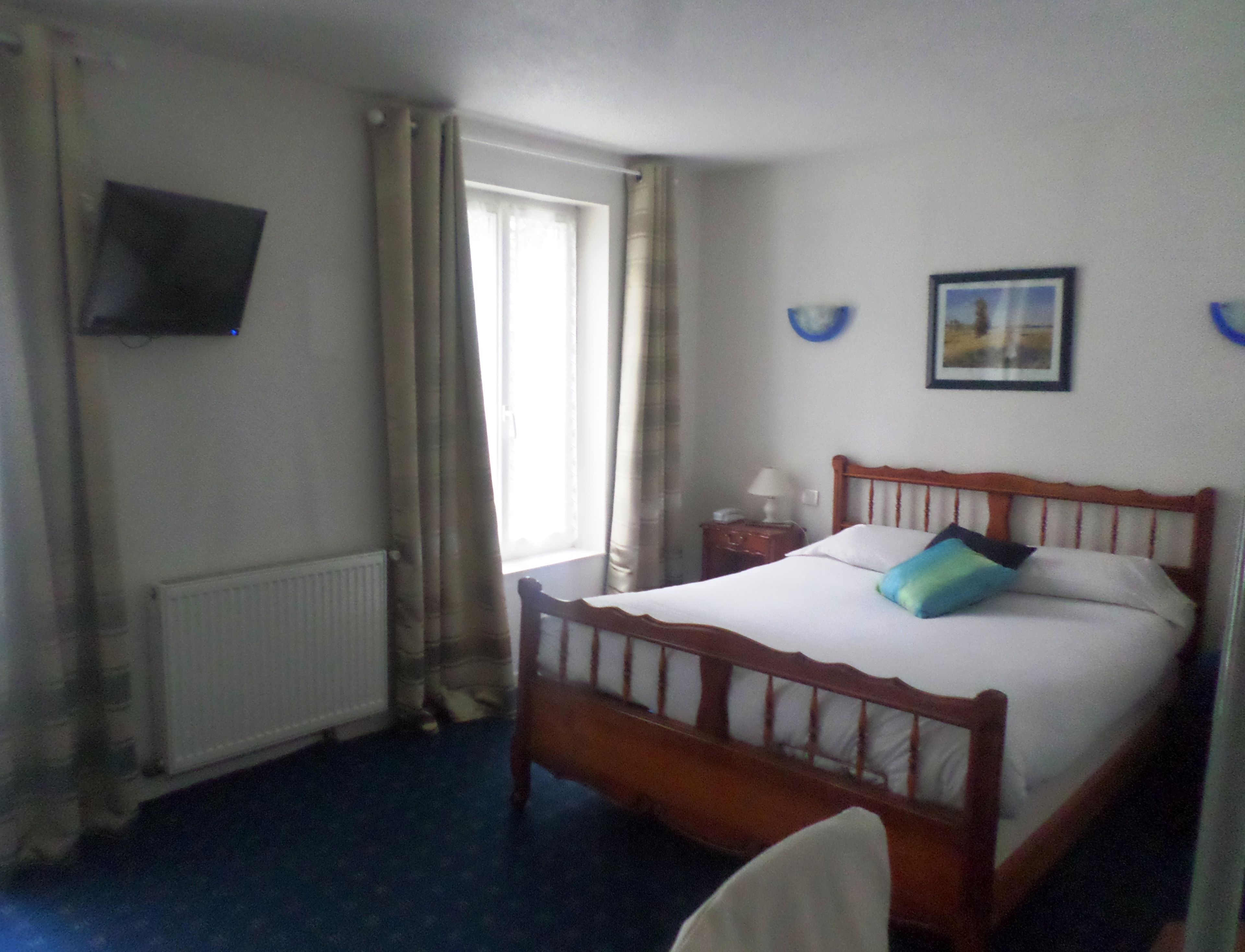 A hotel room with a wooden bed, a nightstand with a lamp, a window with curtains, and a wall-mounted TV.