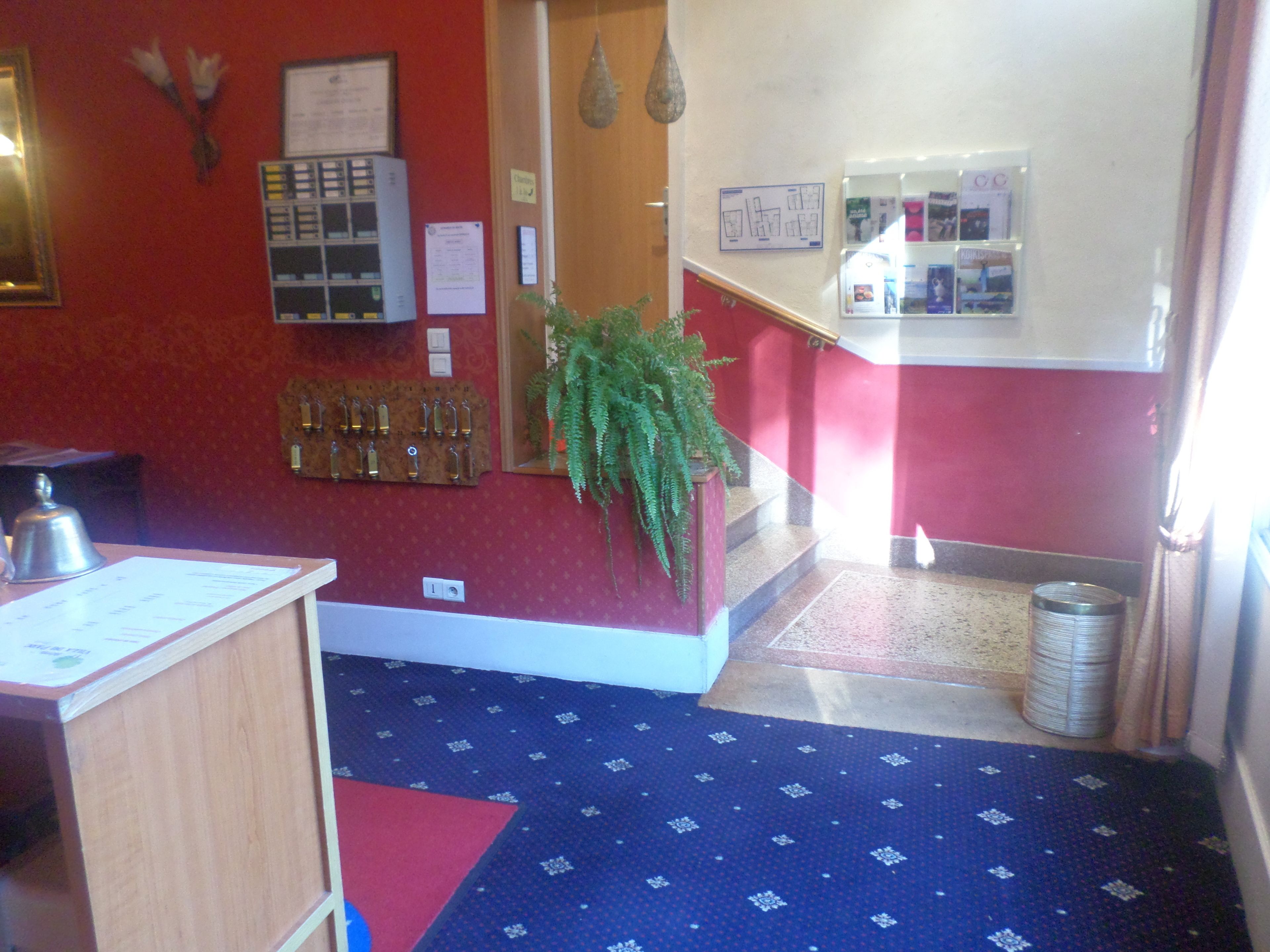 Een hotel lobby met een receptiebalie, een sleutelrek aan de muur, een potplant en een trap naar een andere verdieping.