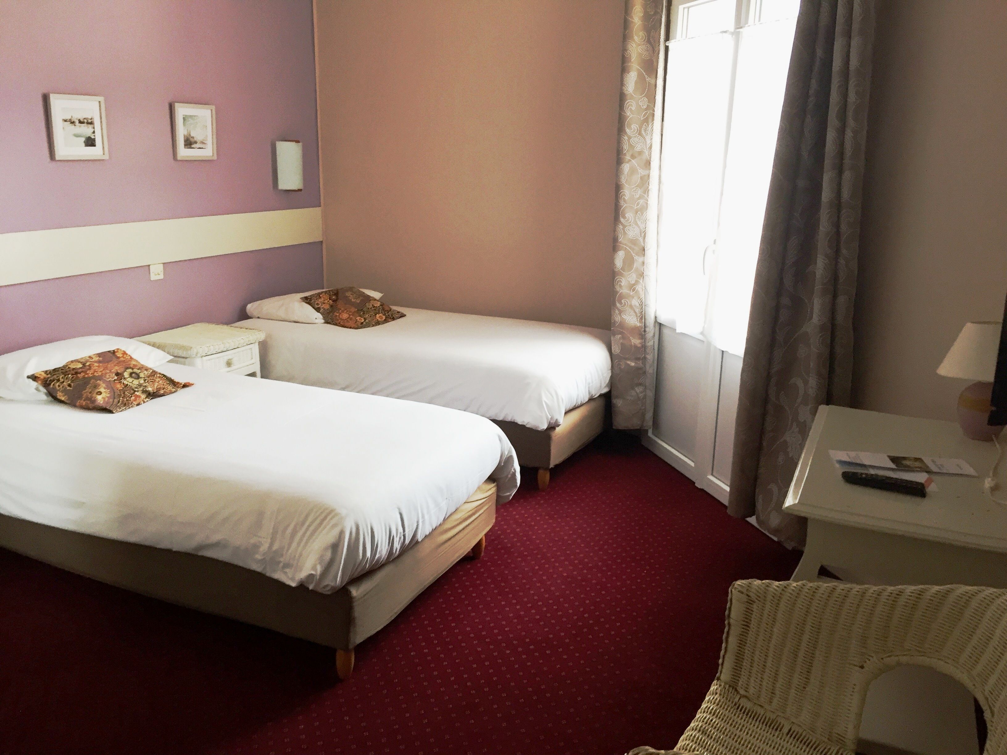 A hotel room with two beds, a red carpet, a white nightstand, and a window with curtains.