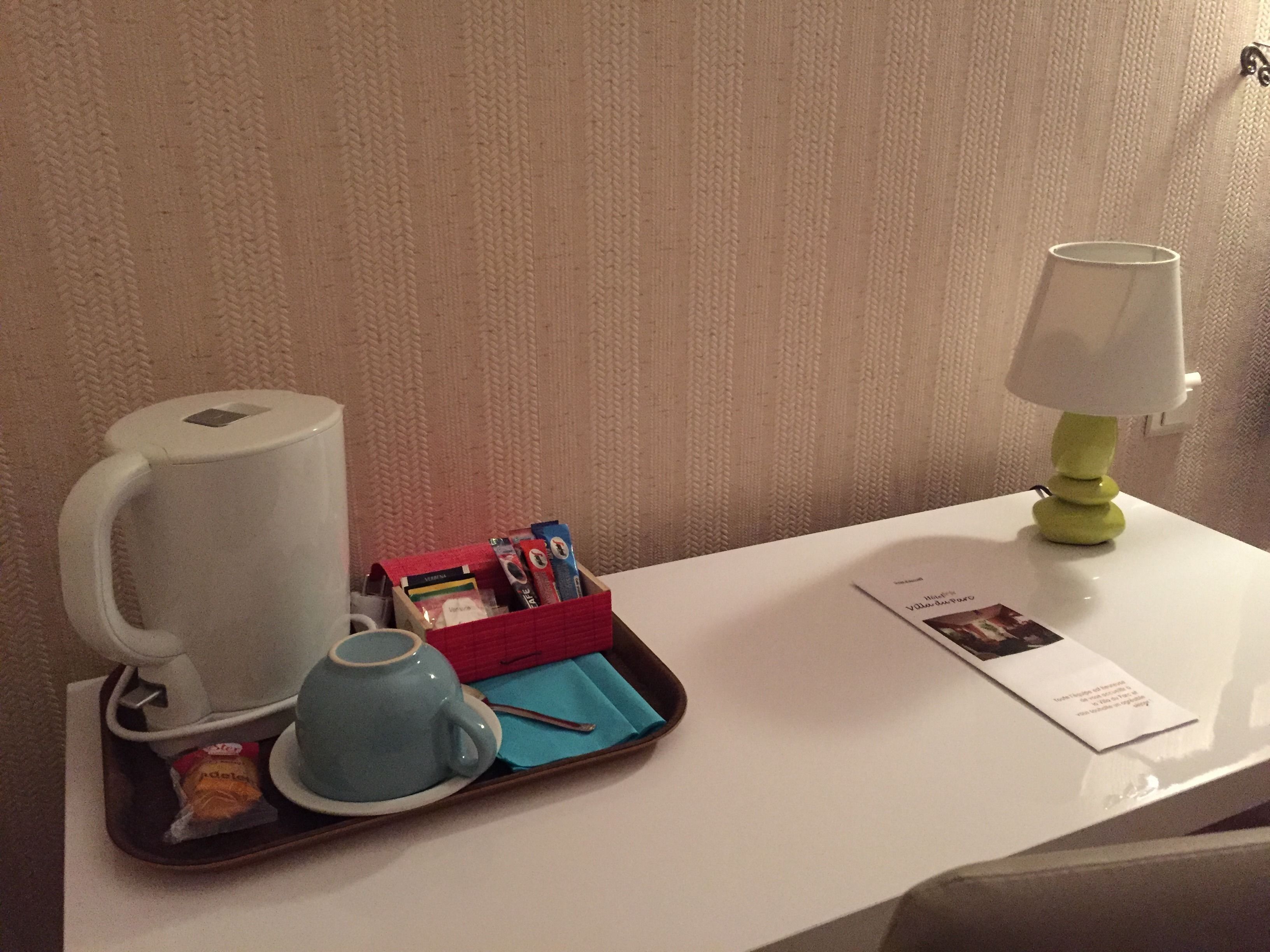 A hotel room with a coffee maker, a tray with tea bags and a spoon, a lamp, and a menu on the table.
