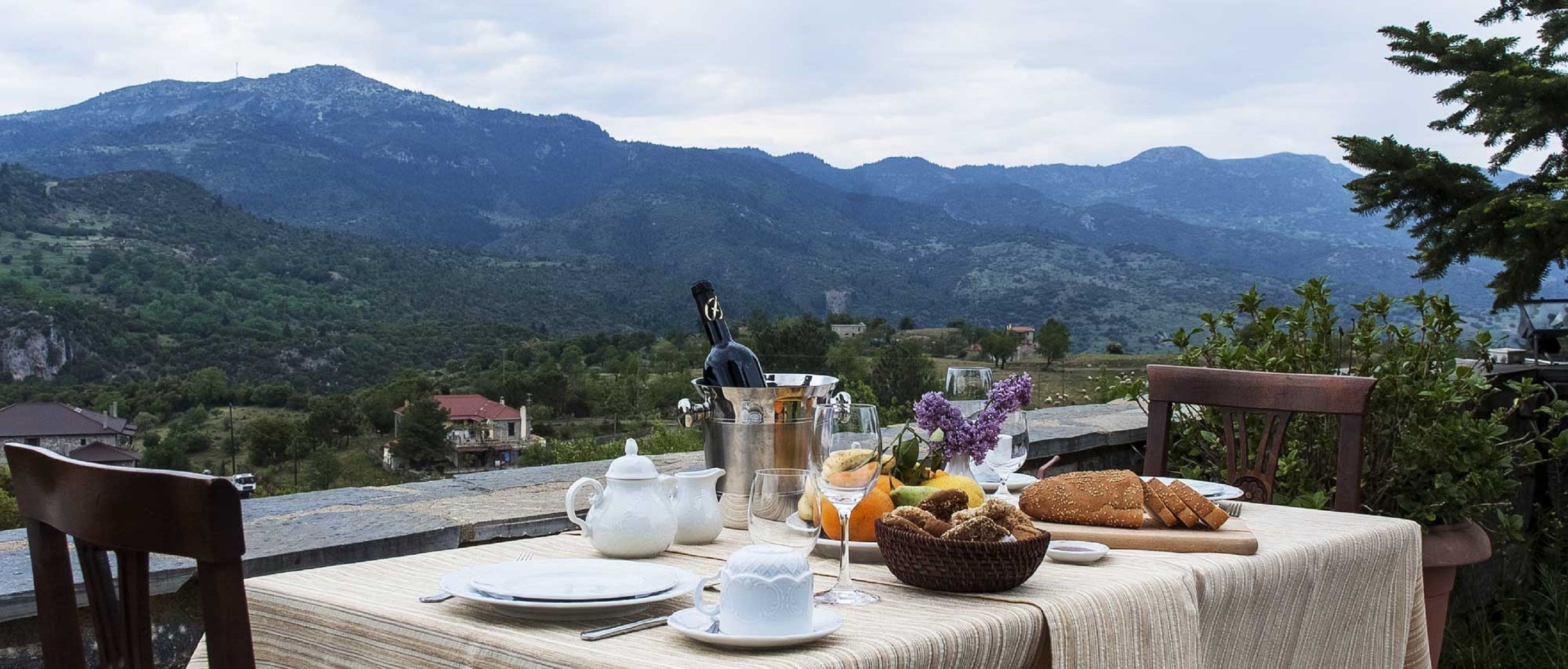 Mesa posta para o café da manhã com vista para a montanha, incluindo um bule, xícaras, pão e frutas.