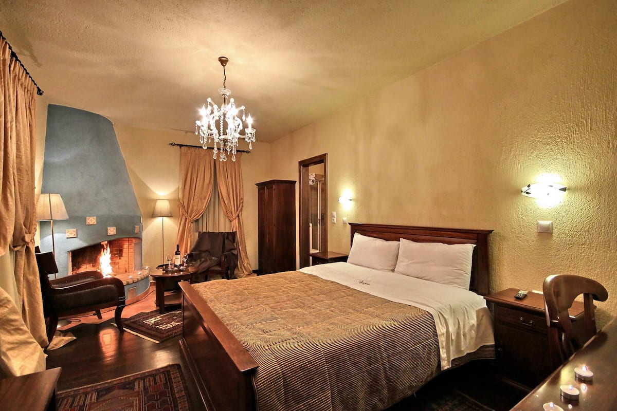 Une chambre d’hôtel avec un grand lit, une table de chevet en bois, une chaise, un bureau, un lustre, une cheminée et un miroir.