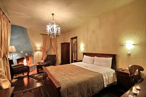 A hotel room with a large bed, a wooden nightstand, a chair, a desk, a chandelier, a fireplace, and a mirror.