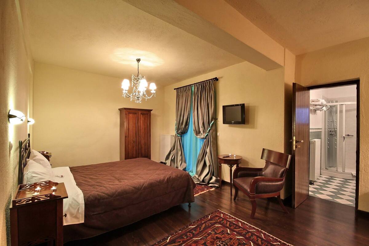 Une chambre d’hôtel avec un grand lit, une table de chevet en bois, une chaise, une petite table, un lustre, une télévision et une salle de bain visible à travers une porte ouverte.