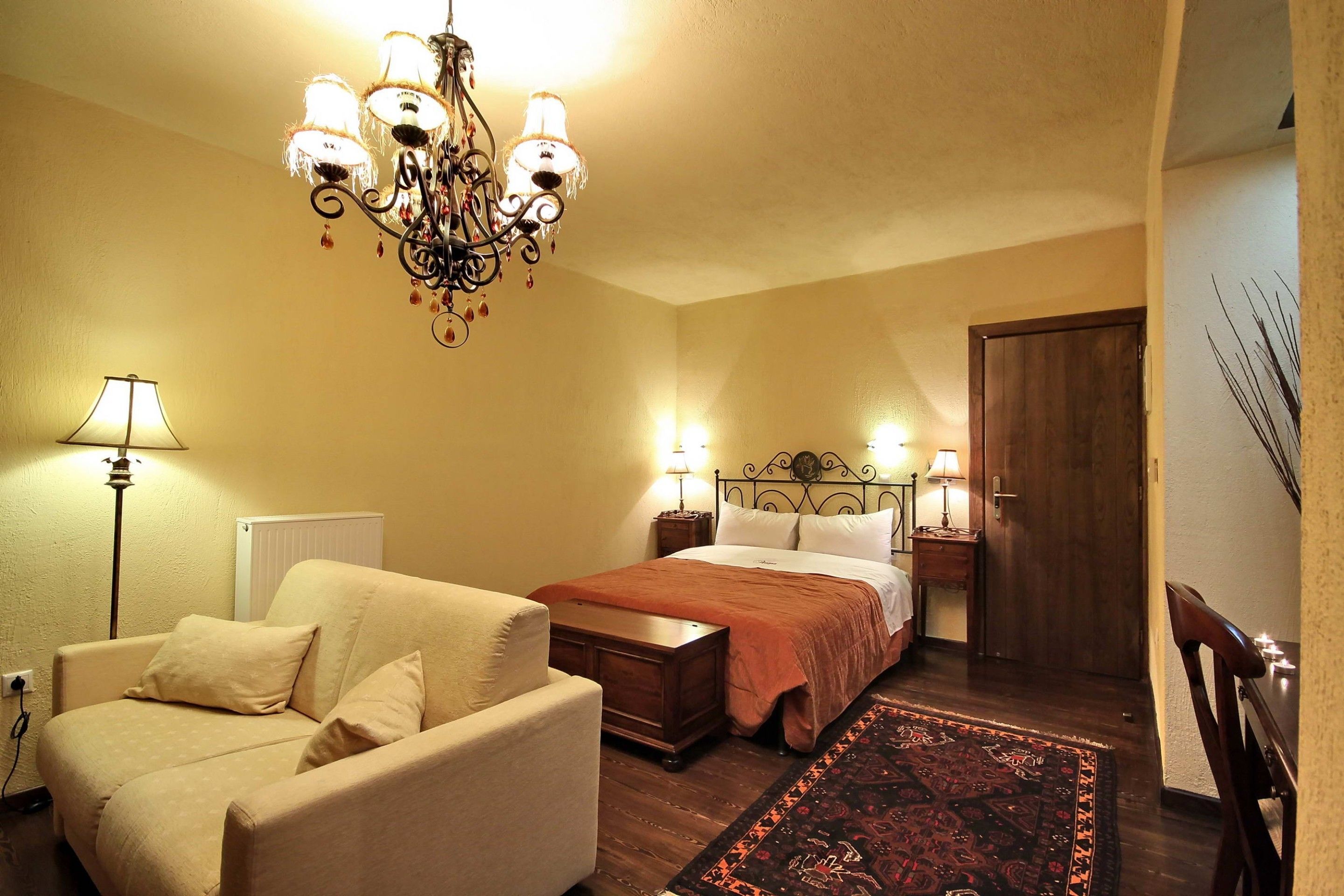 A bedroom with a bed, two bedside tables with lamps, a wooden dresser, a chair, and a chandelier.