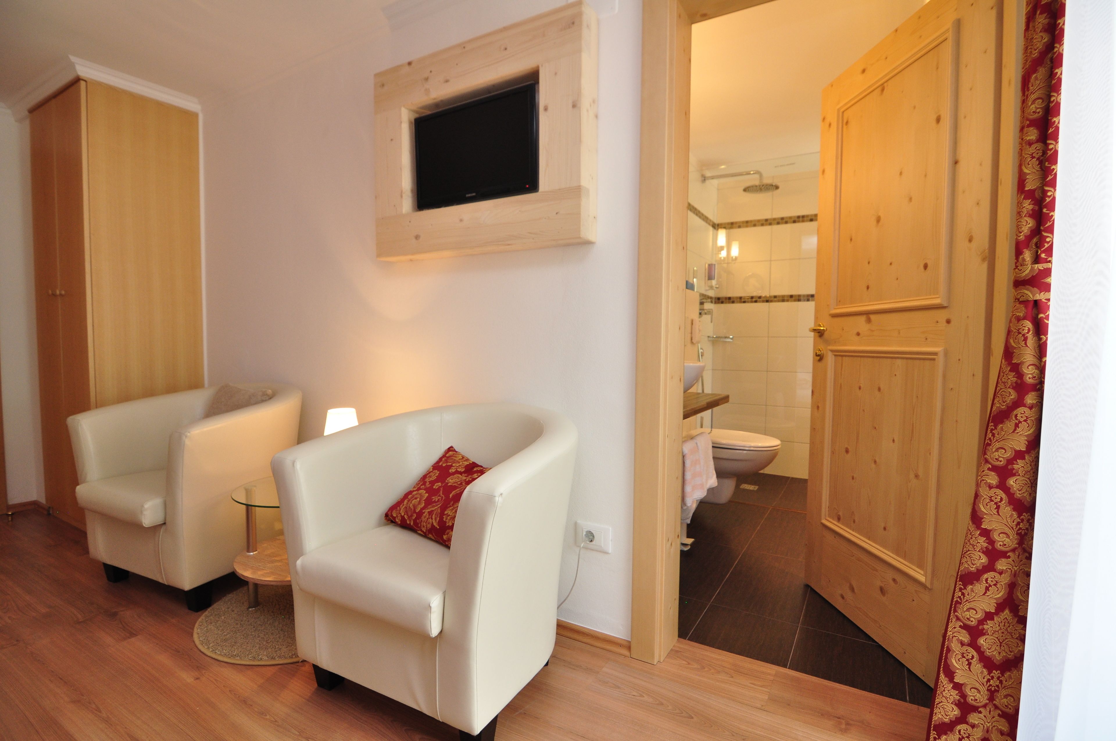 Ein Zimmer mit zwei weißen Sesseln, einem kleinen runden Tisch, einem an einer Holzvertäfelung montierten Fernseher und einer Holztür, die zu einem Badezimmer mit Dusche und Toilette führt.