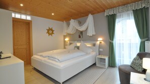A bedroom with a wooden ceiling, a bed with white bedding, a bedside table with a lamp, a nightstand, and a window with green curtains. - Hotel Traube – Stelvio (Stelvio)