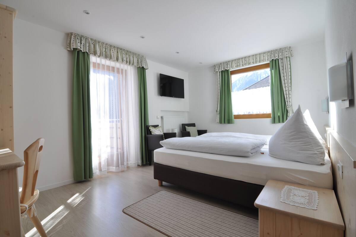 A bedroom with a bed, a desk, a chair, a television, and a window with curtains.