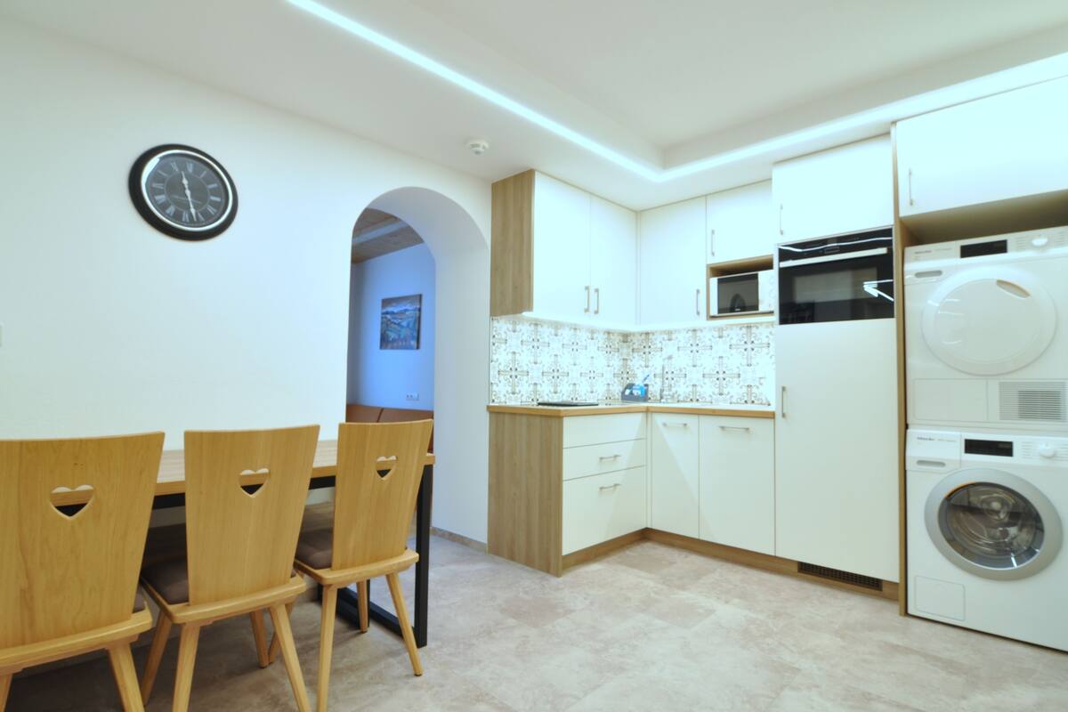 A modern kitchen with white cabinets, a built-in microwave, and a washing machine. There is a dining table with chairs and a clock on the wall.
