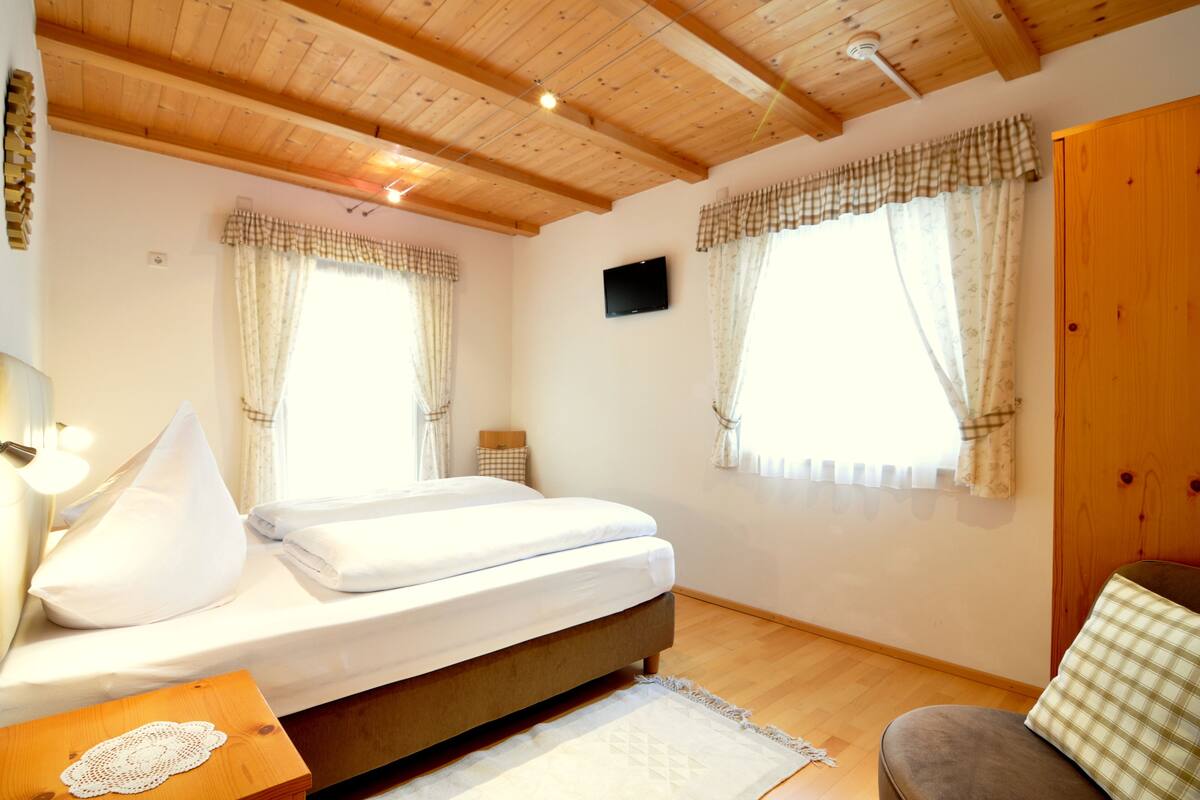 A bedroom with a wooden ceiling, a bed with white linens, two windows with curtains, a television on the wall, and a wooden wardrobe.