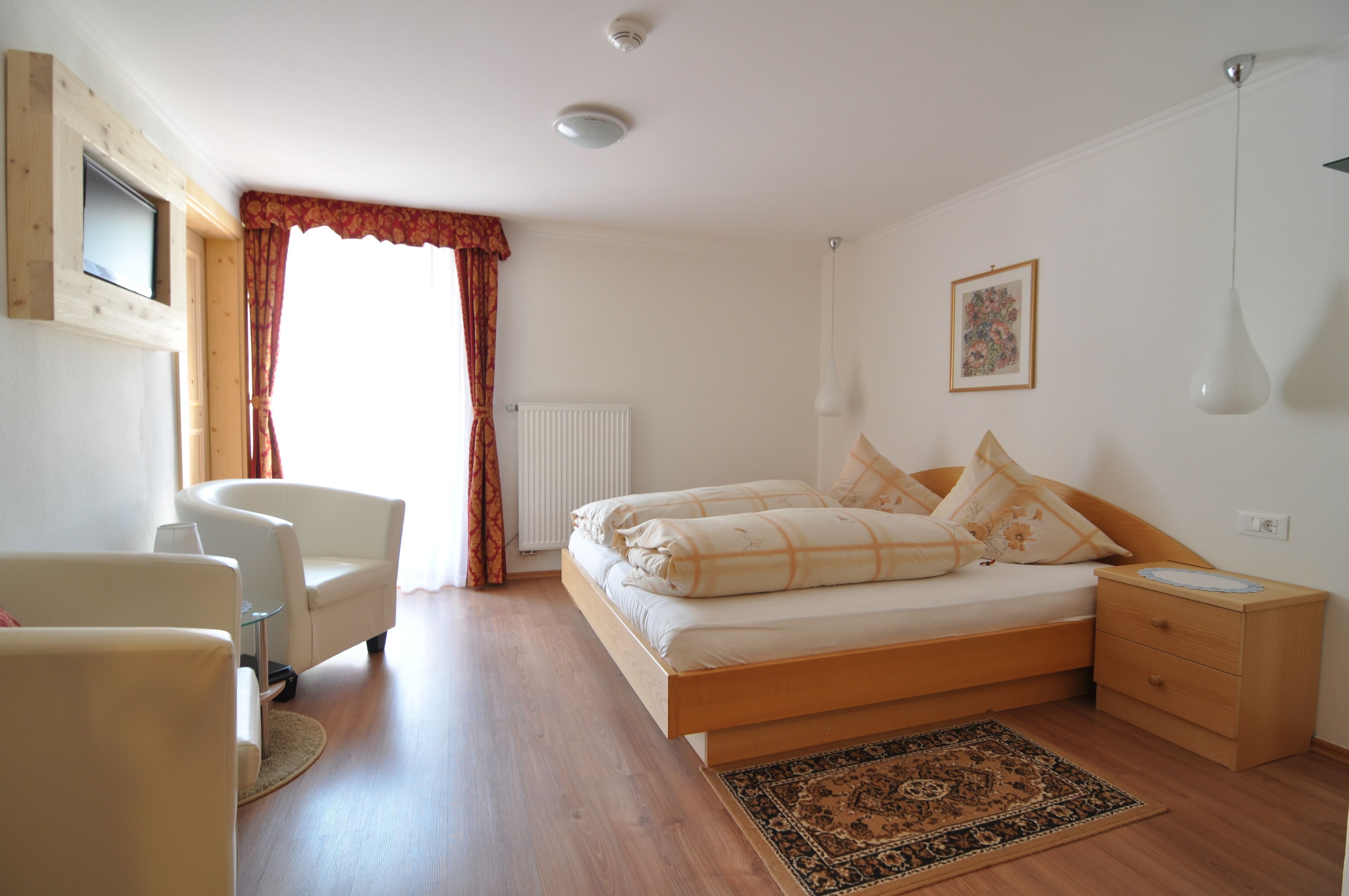 A bedroom with a bed, a chair, a bedside table, a radiator, a window with curtains, and a wall-mounted picture.