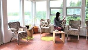 A sunlit room with wicker chairs, a table, and large windows offering a view of greenery. -  Veghús Guesthouse (Selfoss)