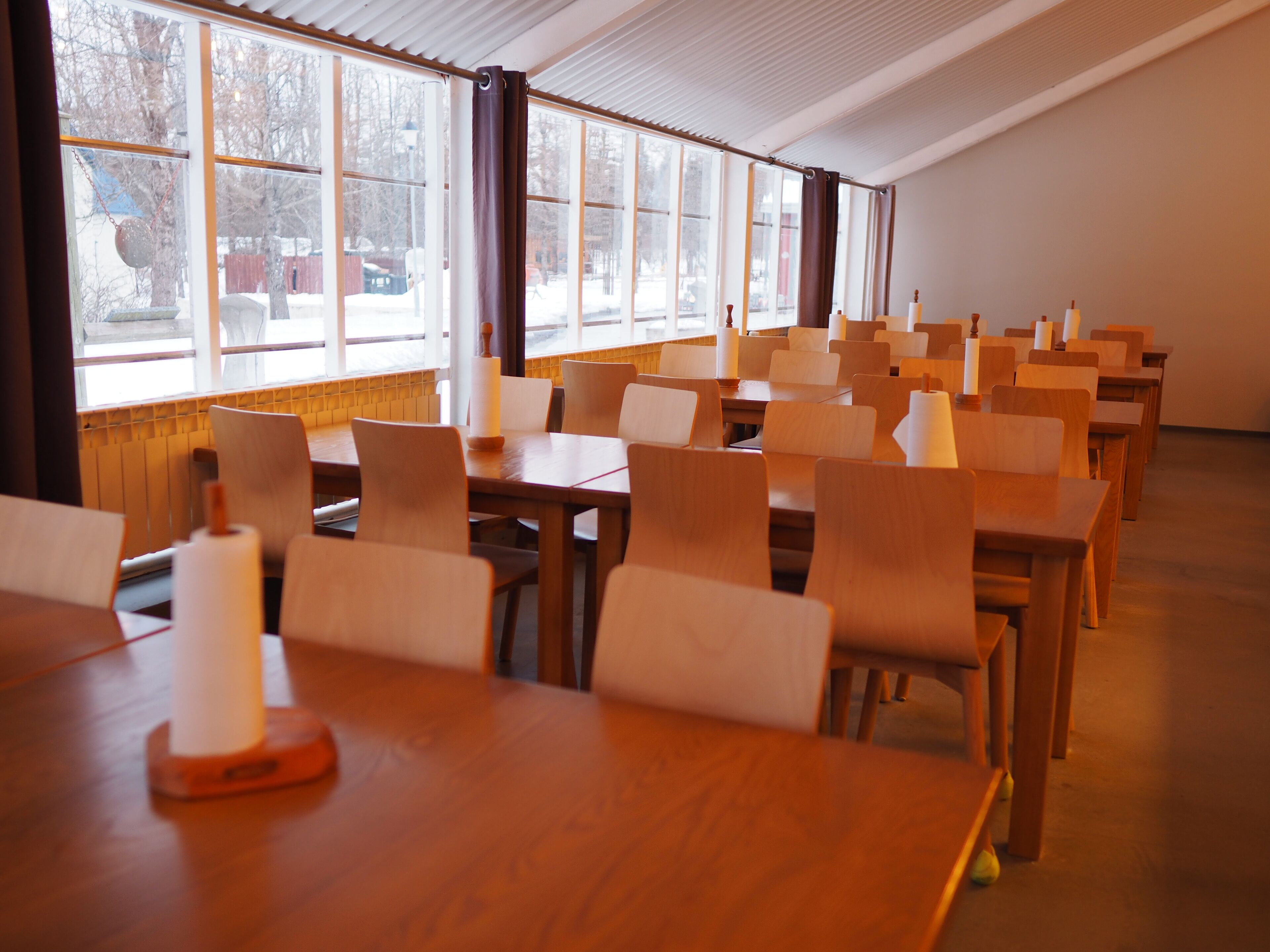 A dining area with wooden tables and chairs, large windows, and a view of trees outside.