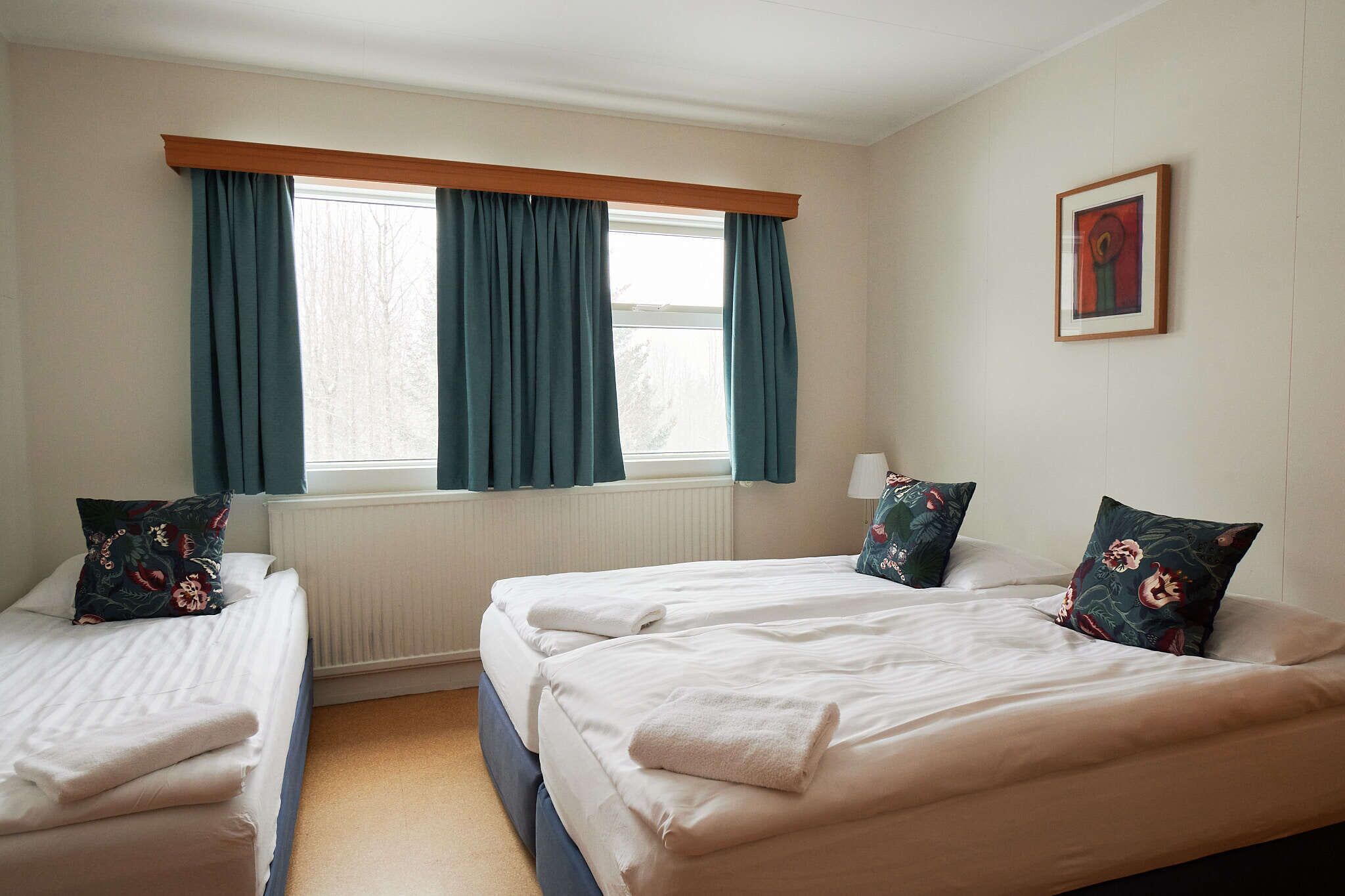 Een kamer met twee bedden, een raam met gordijnen, een radiator en een lijst met een afbeelding aan de muur.