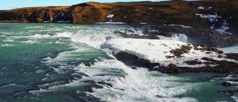 Paisaje costero con olas turbulentas rompiendo contra costas rocosas, parches de nieve en las colinas y un cielo despejado.