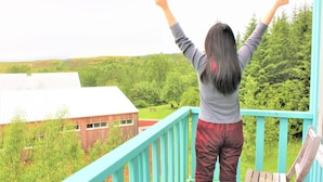 A person standing on a balcony with arms raised, overlooking a green landscape with buildings in the background. -  Veghús Guesthouse (Selfoss)