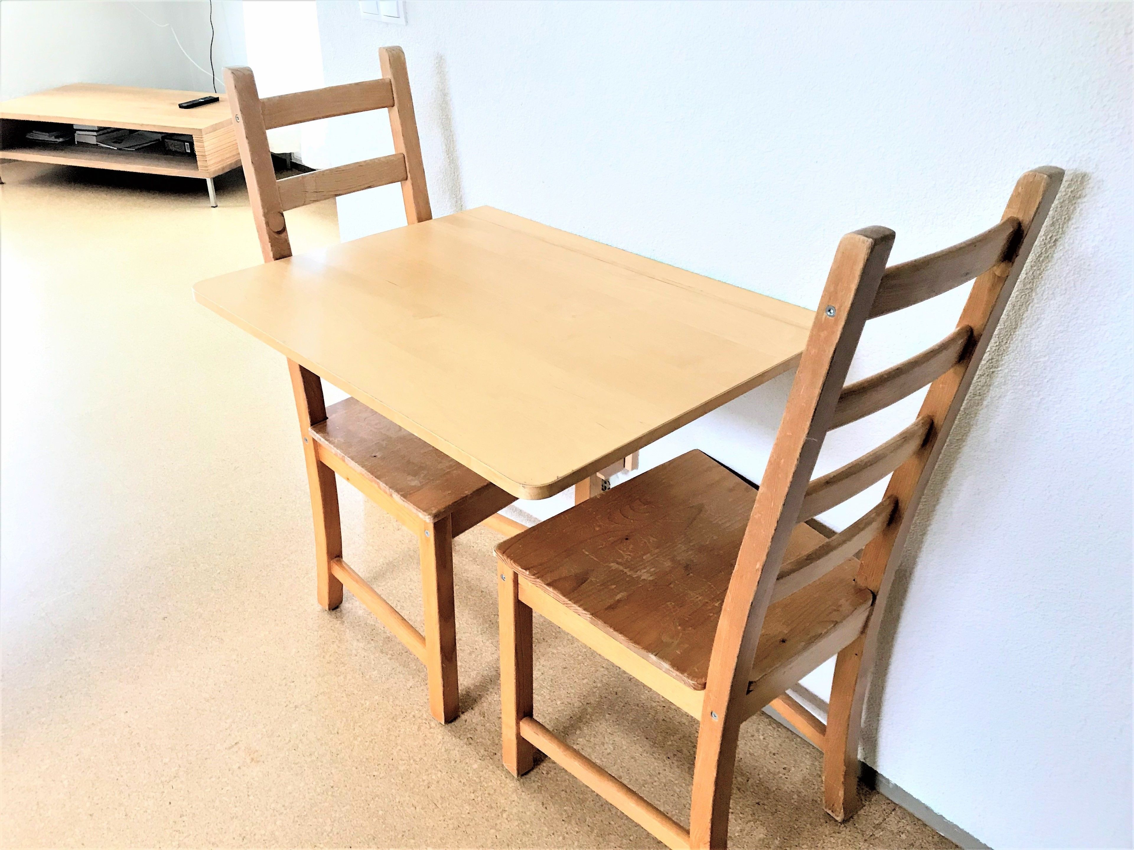 Una mesa de comedor de madera con cuatro sillas, un estante de madera pequeño y un control remoto sobre la mesa.