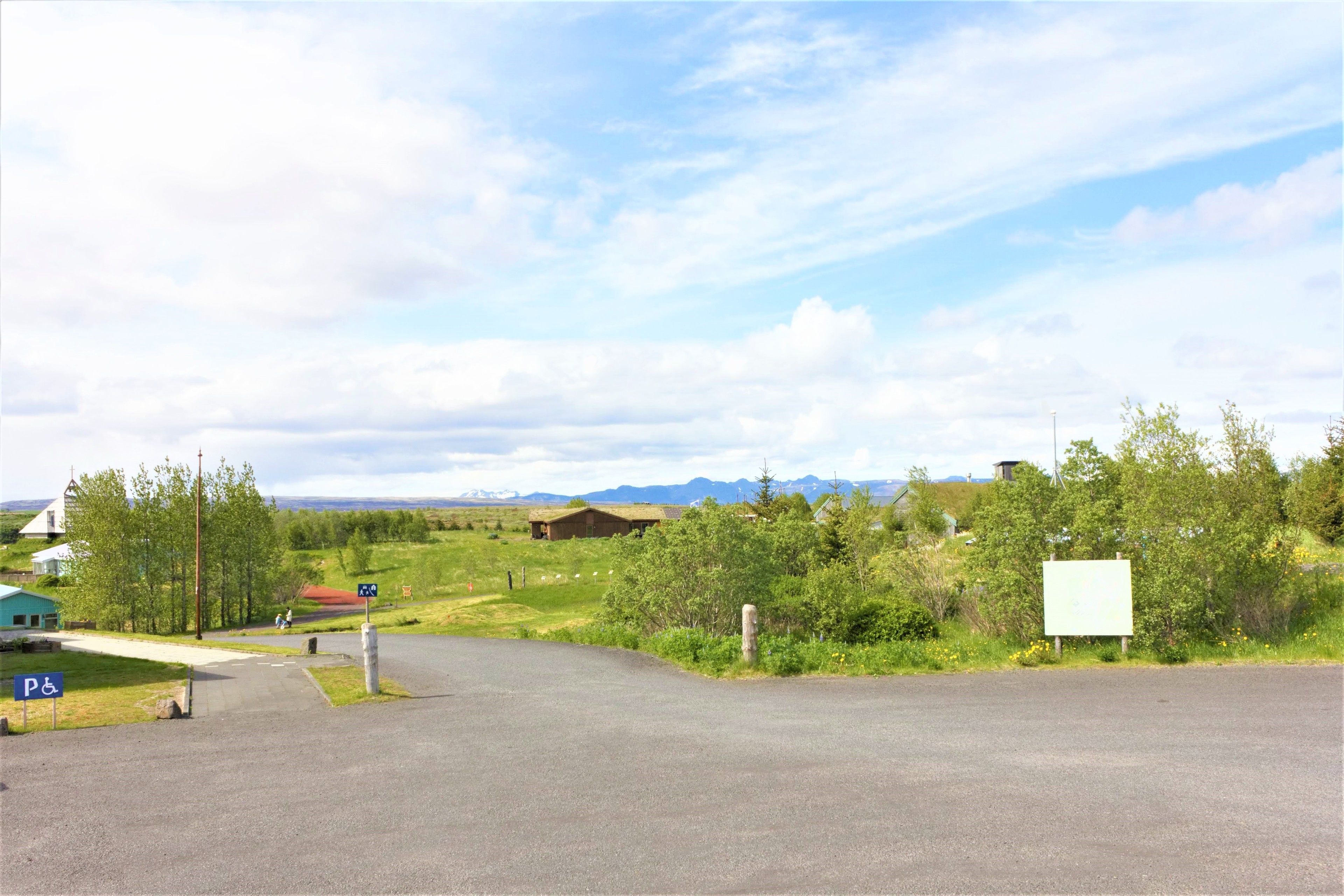 A parking area with a wheelchair accessible sign, a building with a thatched roof, and a mountain range in the background.