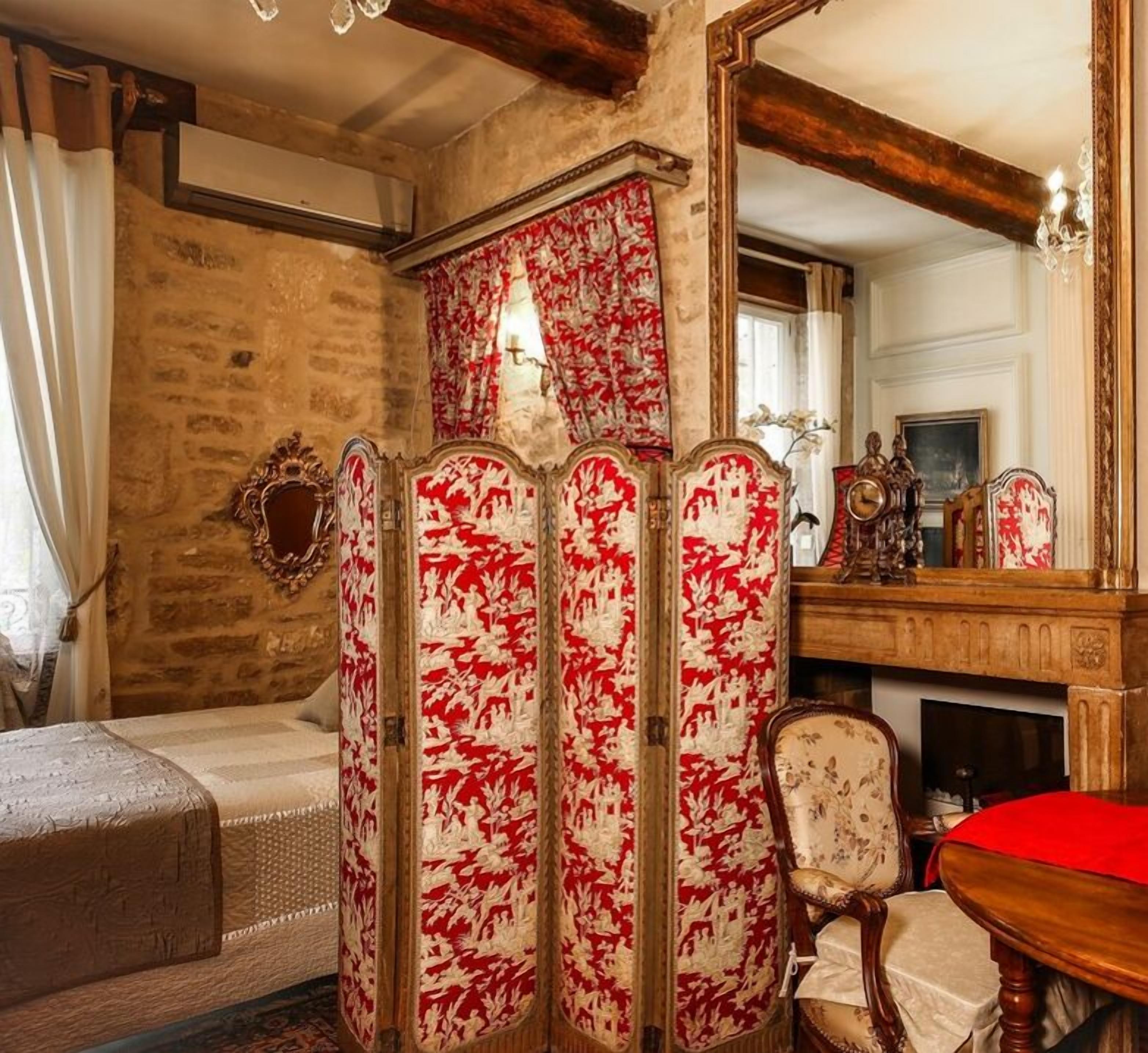A traditional room with a stone wall, a bed, a red and white patterned screen, a wooden chair, and a fireplace.