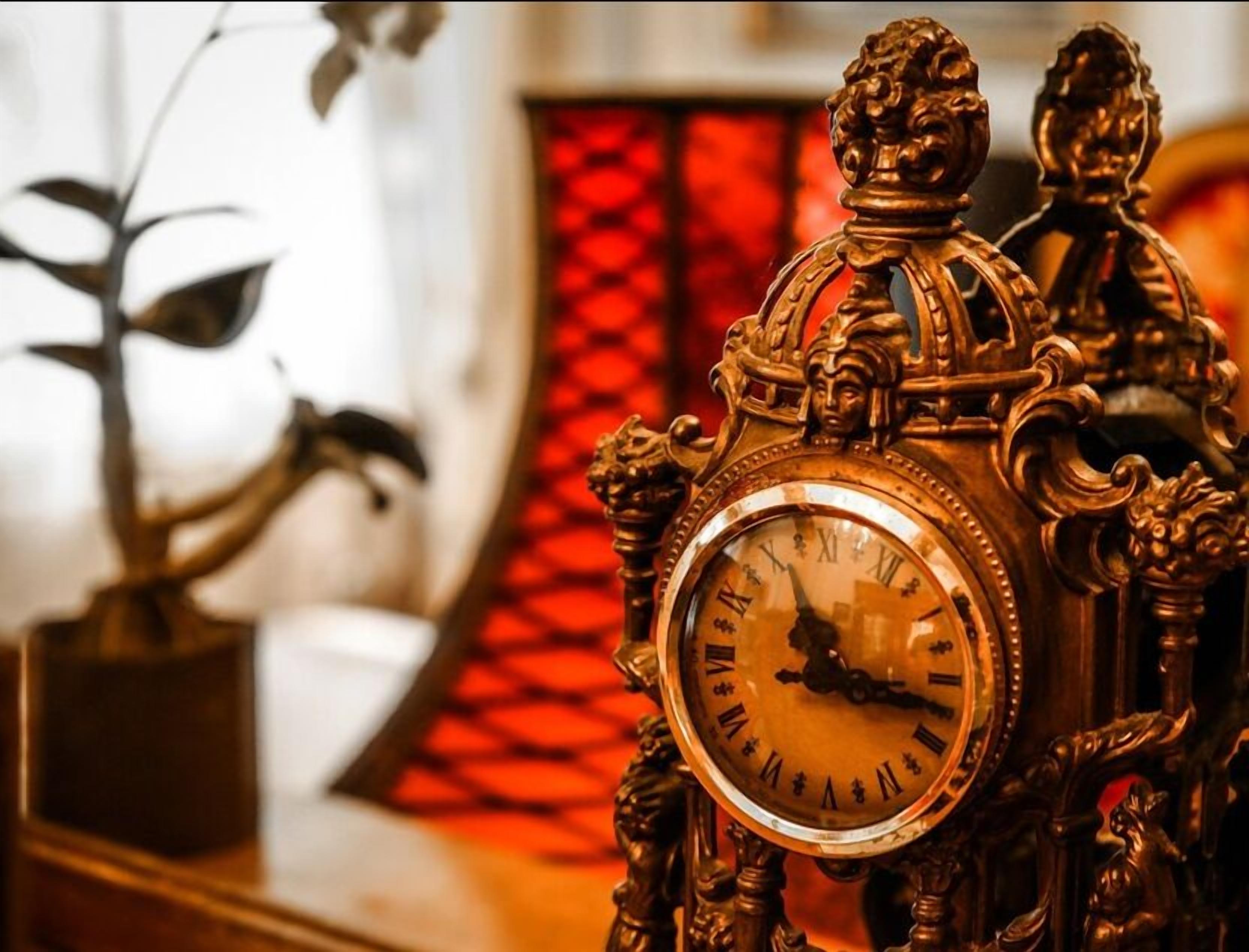 An ornate clock with Roman numerals, set against a blurred background with a red patterned wall.
