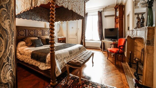 A four-poster bed with a lace canopy, a television on a wooden stand, and a red armchair in a room with wooden flooring and a patterned rug. - Le Petit Tertre (Dijon)