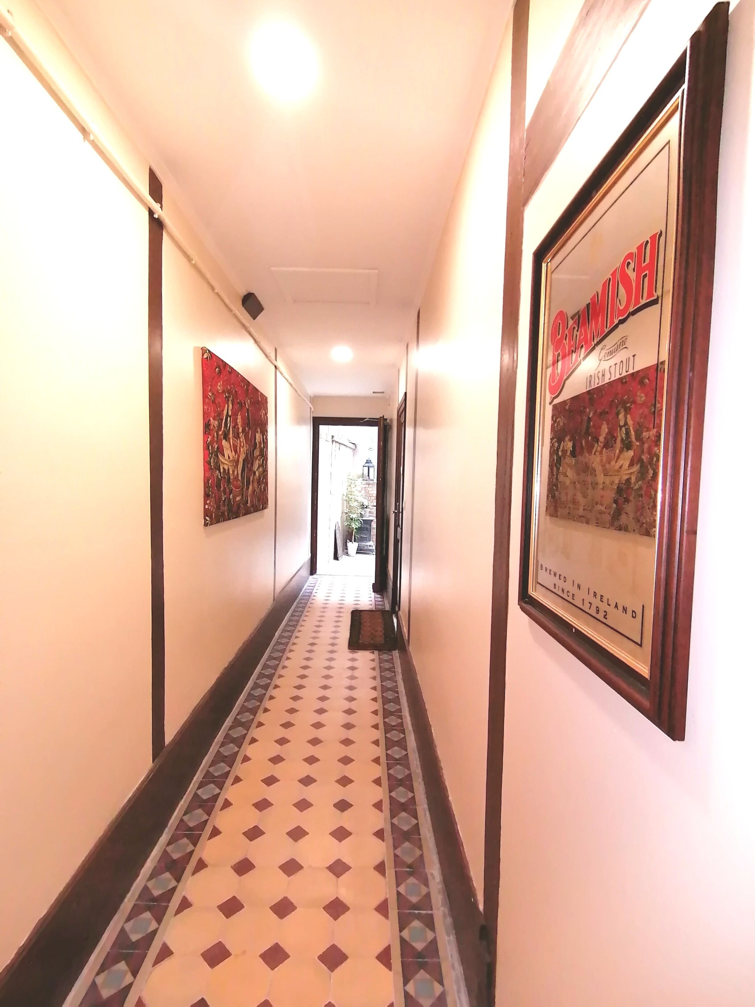 Ein Hotelkorridor mit gemusterten Bodenfliesen, gerahmten Kunstwerken an den Wänden und einem gerahmten Werbeplakat einer Biermarke.
