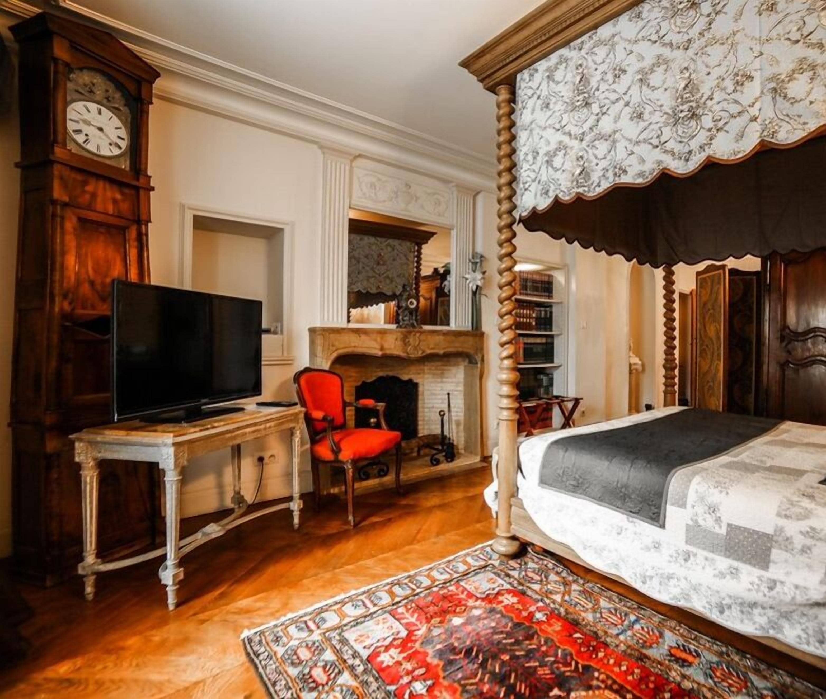 Une chambre à coucher avec un lit à baldaquin, un téléviseur posé sur un bureau en bois, un fauteuil rouge, une cheminée et une grande horloge.