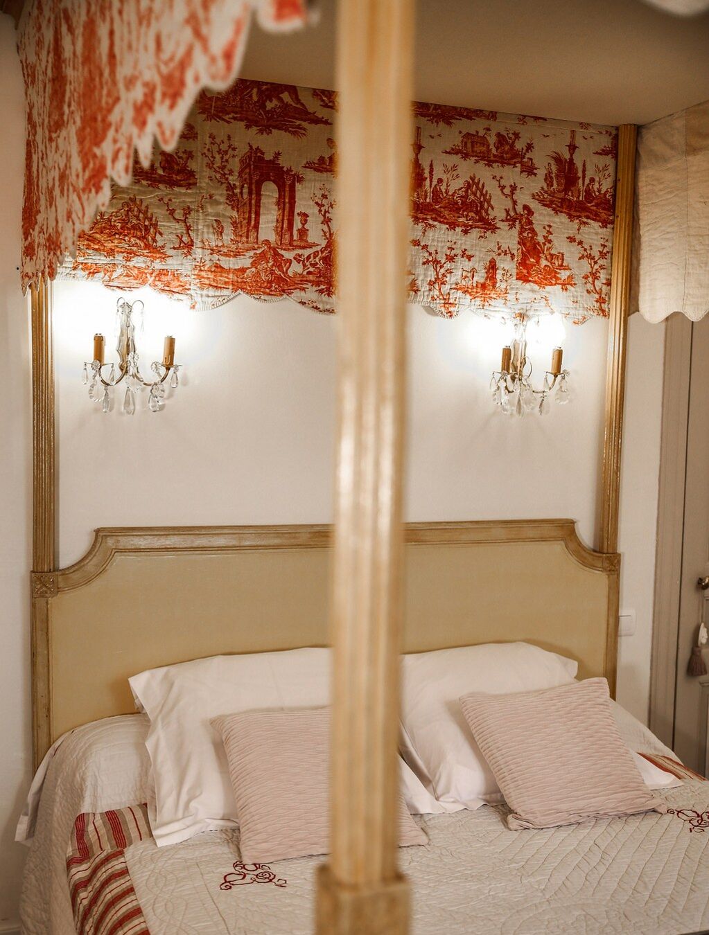 A bedroom with a four-poster bed, two wall sconces, and a patterned tapestry.