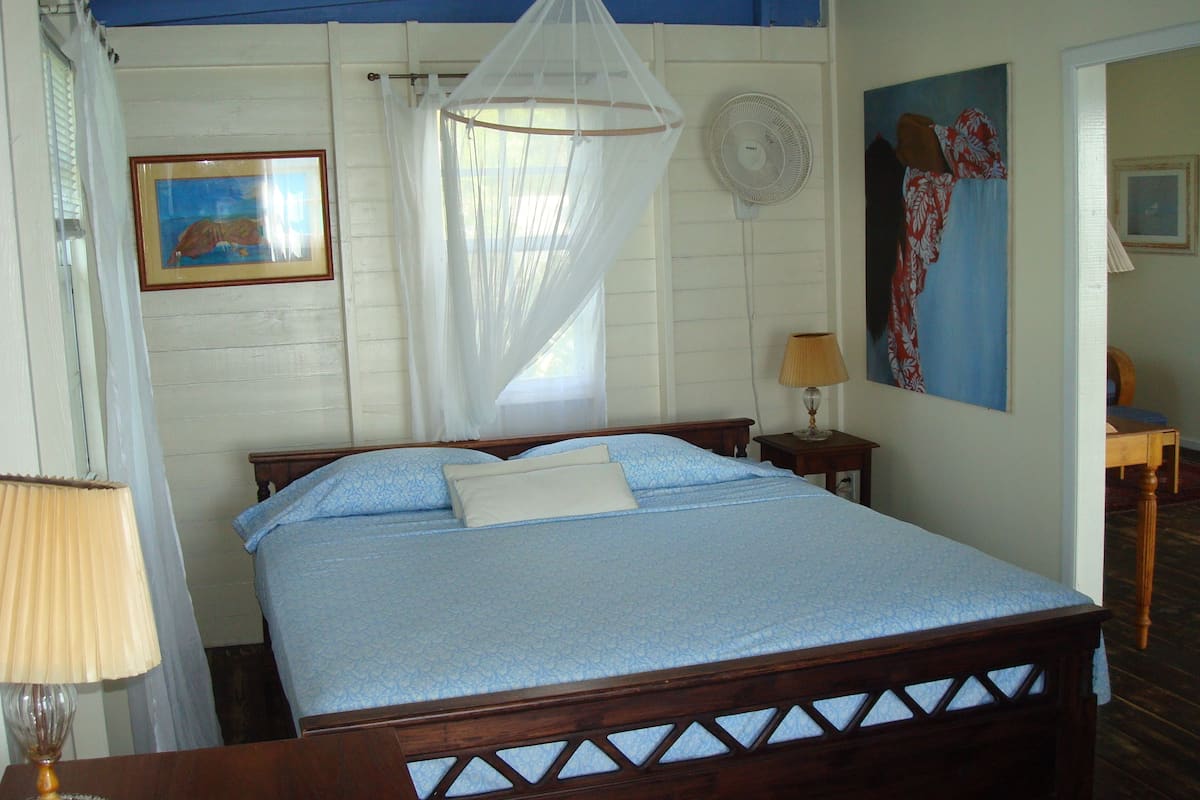 A bedroom with a bed, a bedside table with a lamp, a standing fan, and a painting on the wall.