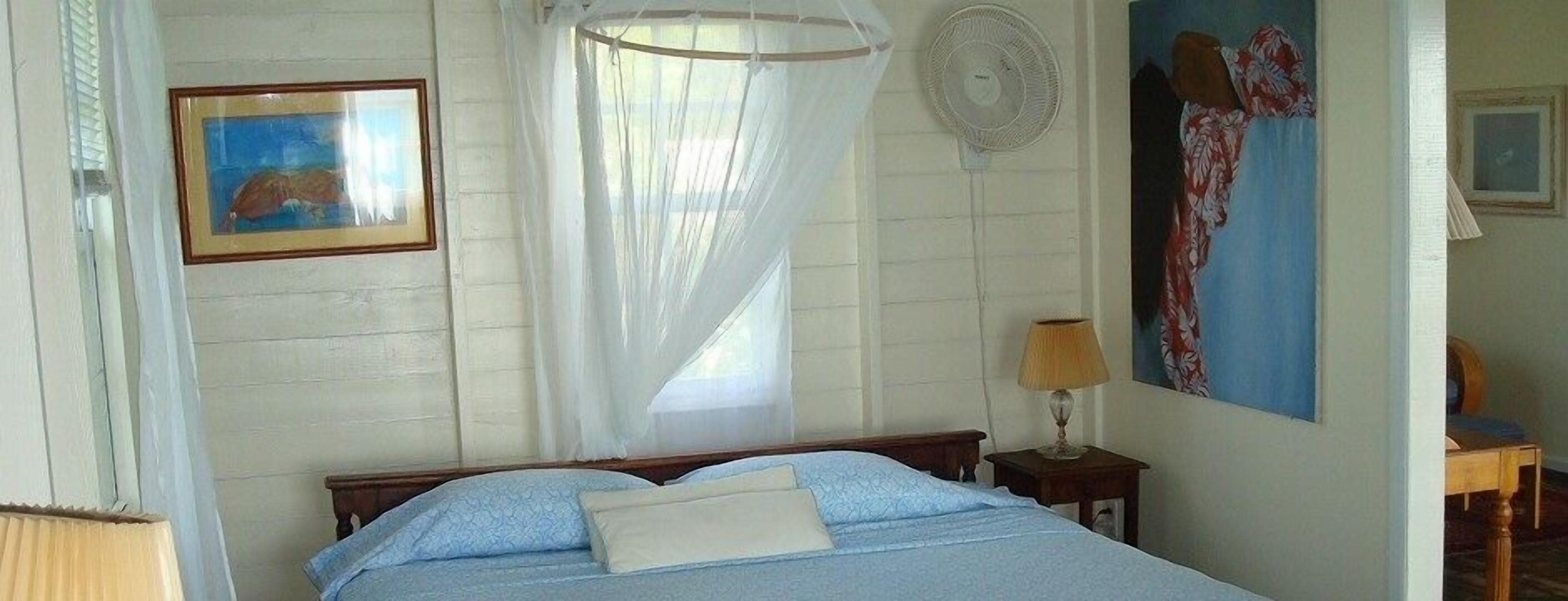 A bedroom with a bed, a bedside table with a lamp, a standing fan, a window with curtains, and a painting on the wall.