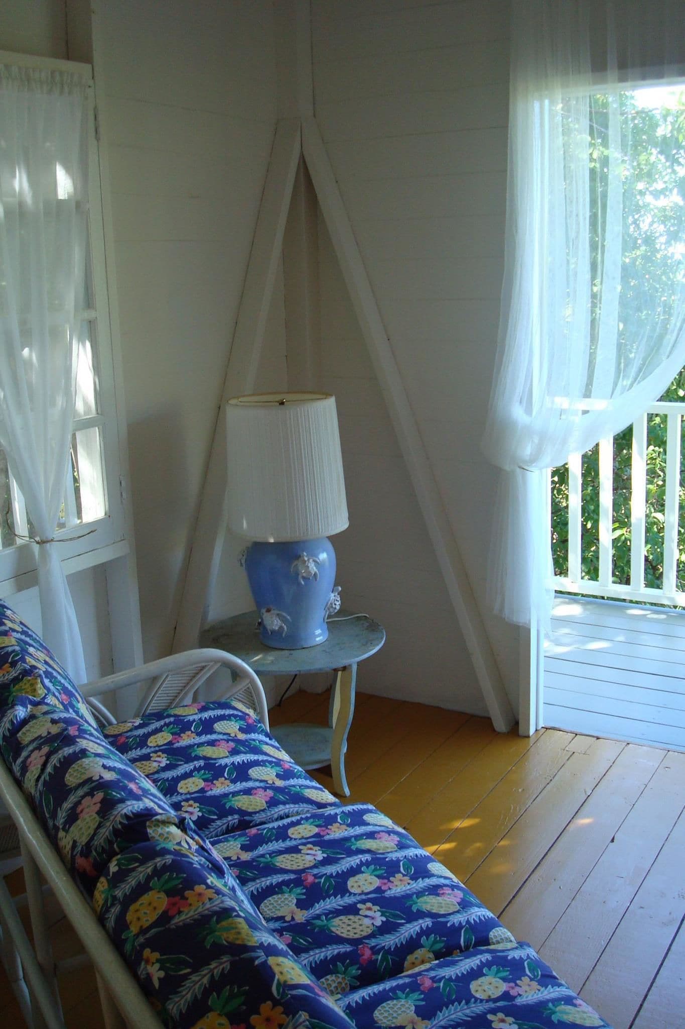 A room with a wooden floor, a bed with a floral-patterned bedspread, a small table with a lamp, and a window with sheer curtains.