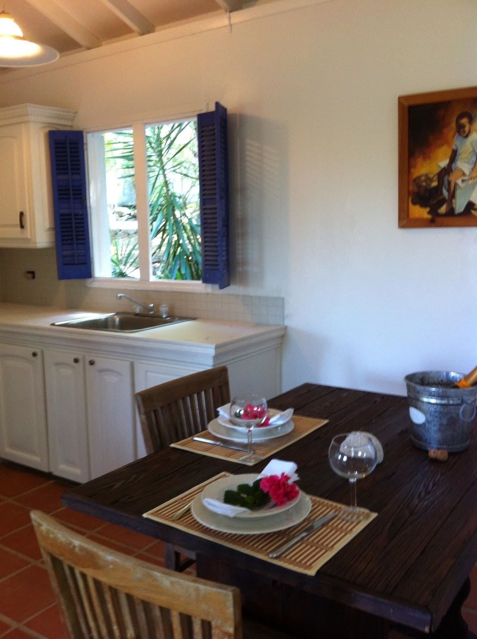 A dining area with a wooden table set for two, a window with blue shutters, and a kitchen in the background.