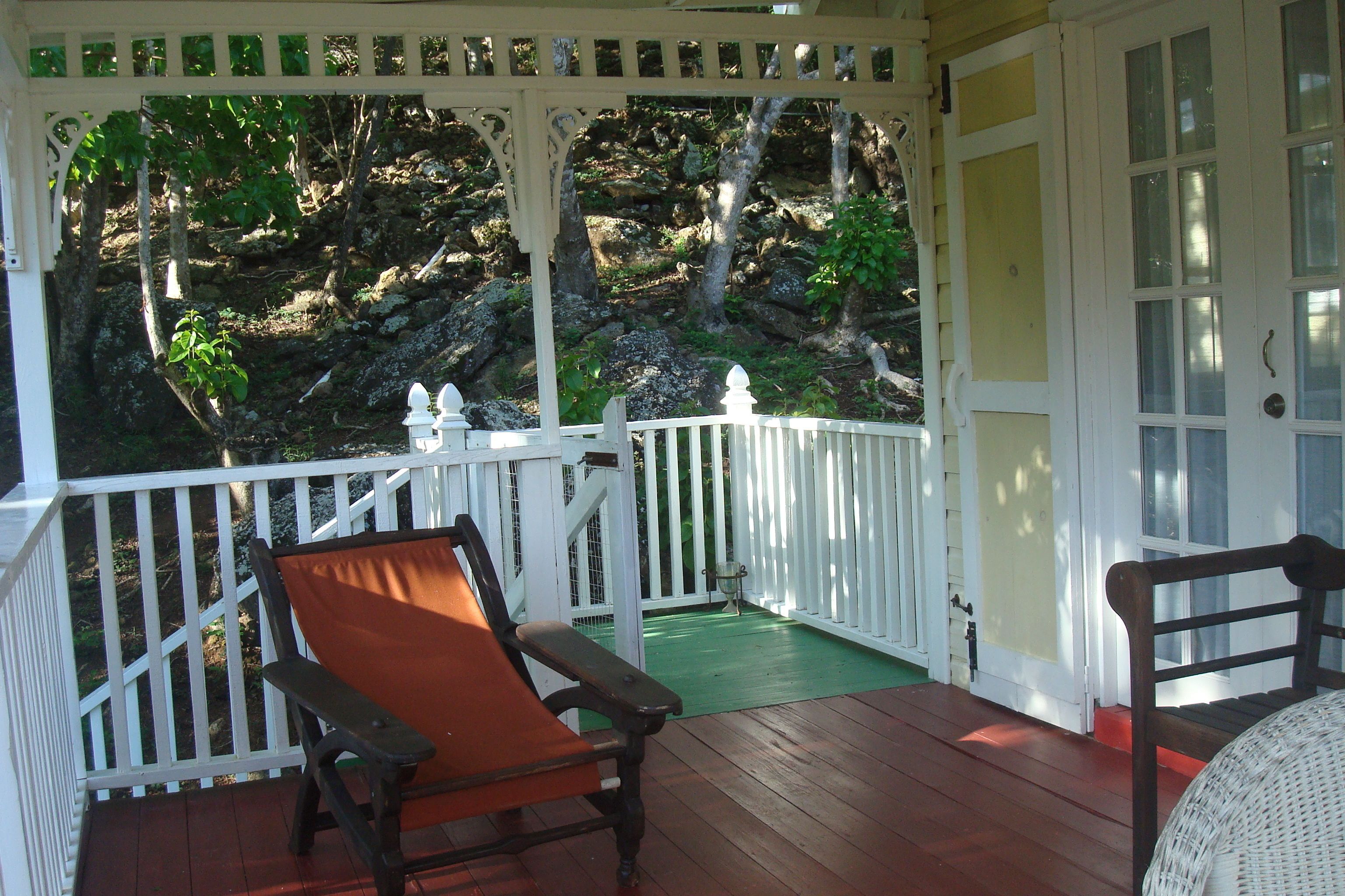 A wooden deck with a white railing, a bench, and a door leading to a green area with a rocky backdrop.