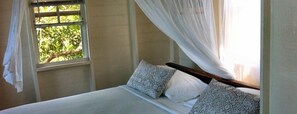 Cama com roupa de cama branca e almofadas cinzentas, janela com vista para vegetação e cortina branca.