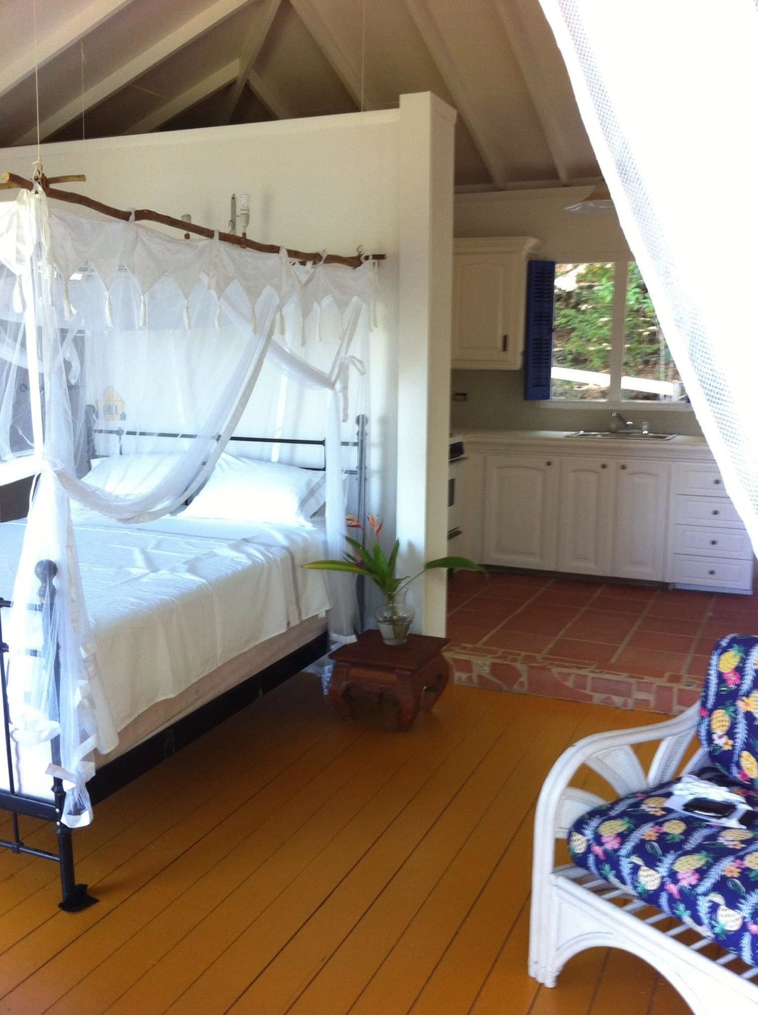 A bedroom with a canopy bed, a small table with a plant, a white chair, and a view of a kitchen and outdoor area.