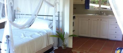 A bedroom with a canopy bed, a small table with a plant, a white chair, and a view of a kitchen and outdoor area.