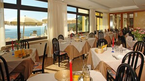 A restaurant with multiple tables set for dining, large windows with curtains, and a view of the sea. - Hotel Residence La Rotonda (Tignale)