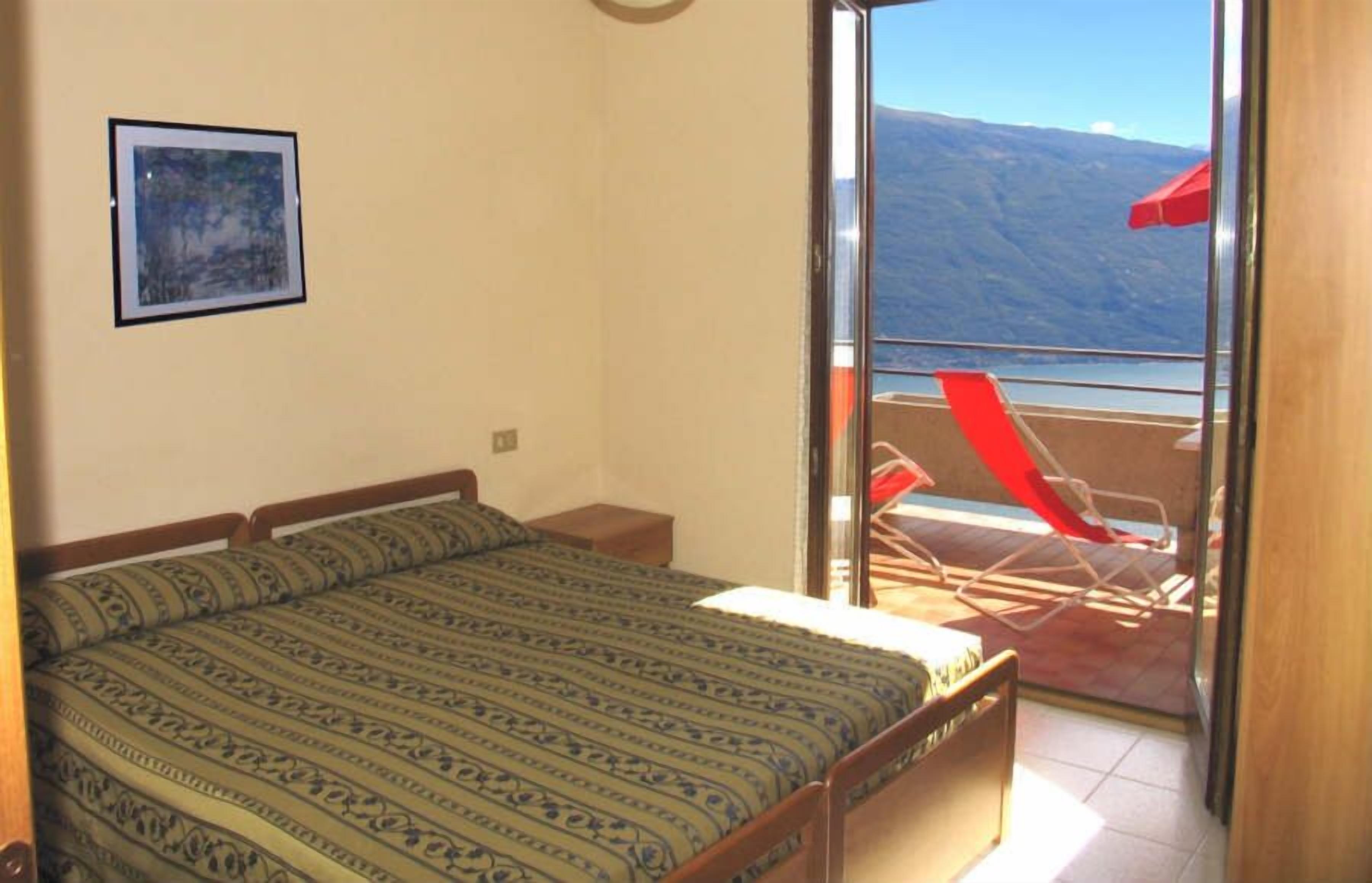 Un dormitorio con cama, una mesita de noche pequeña y un cuadro en la pared. Cuenta con un balcón con una silla y un paraguas rojos.