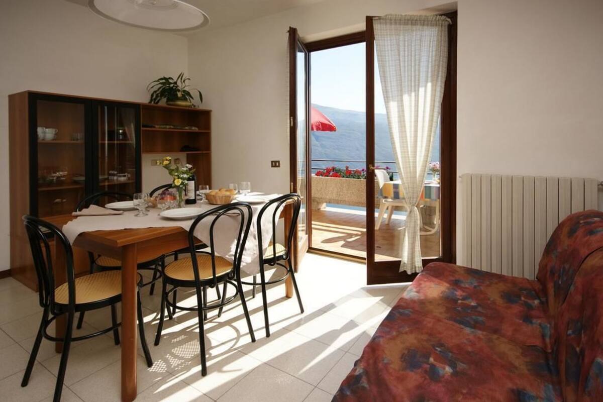 A room with a dining table set for four, a sofa, a bookshelf, and a view of the sea through an open door.