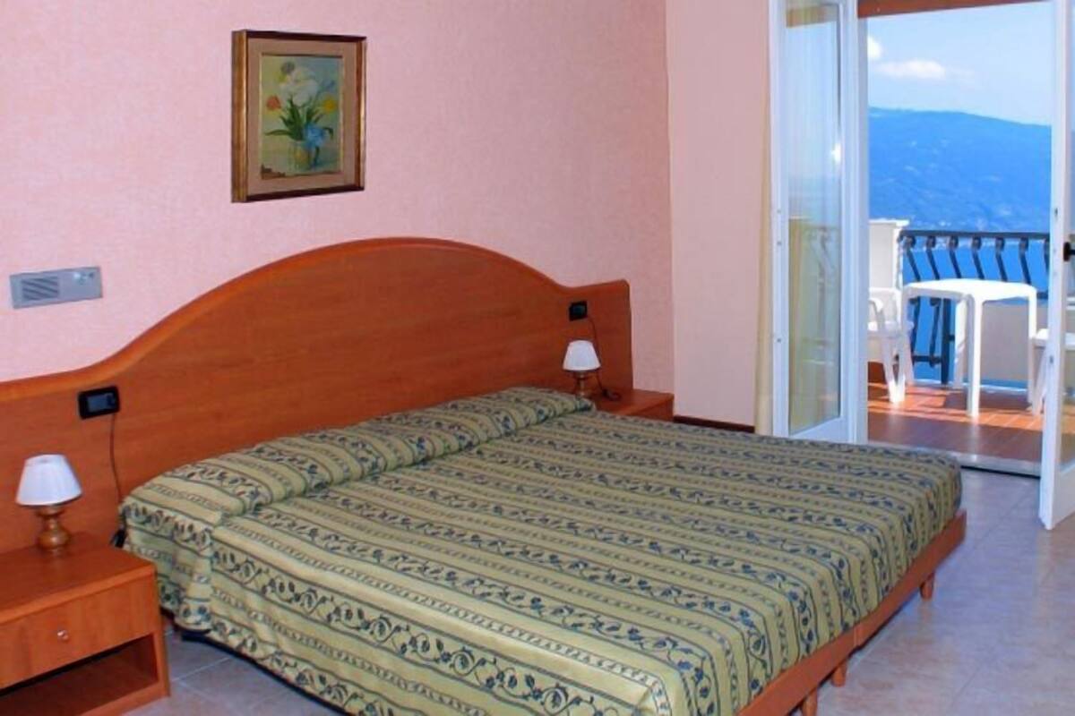 A hotel room with a bed, bedside lamps, a TV, and a balcony with a view of the sea.