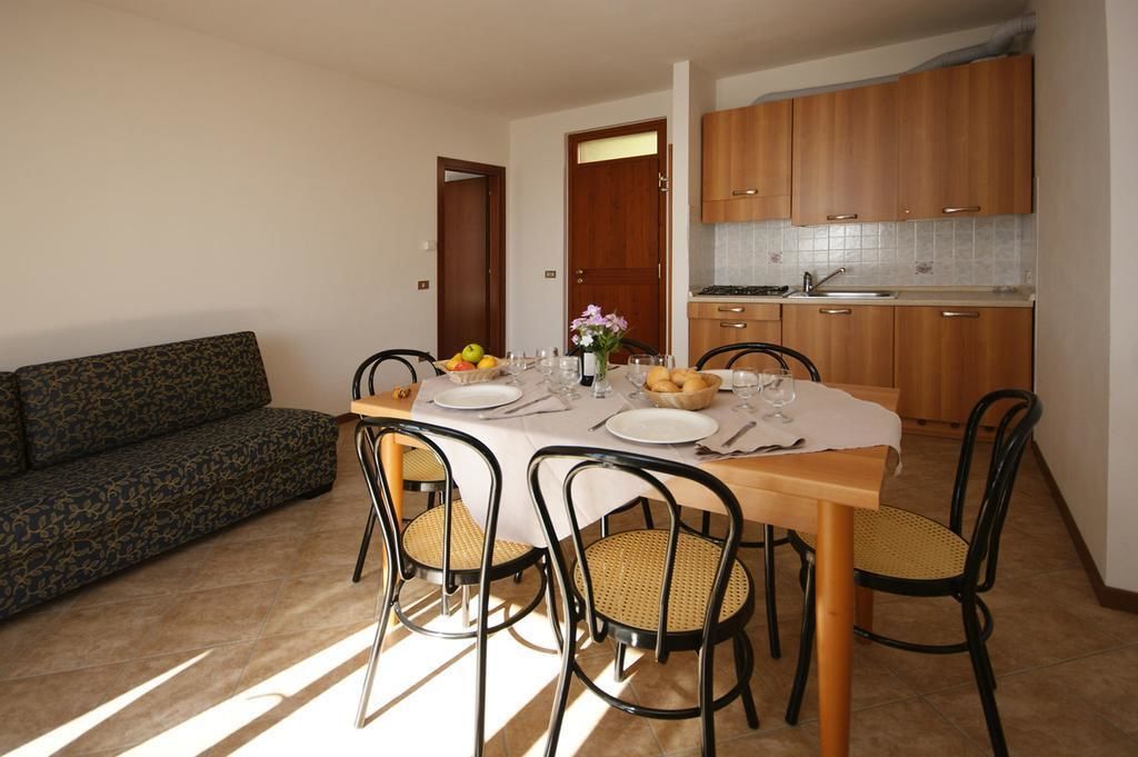 Zona pranzo con tavolo in legno apparecchiato per quattro persone, circondato da sedie in vimini. Sullo sfondo si vede un divano, e una cucina con pensili in legno è adiacente.